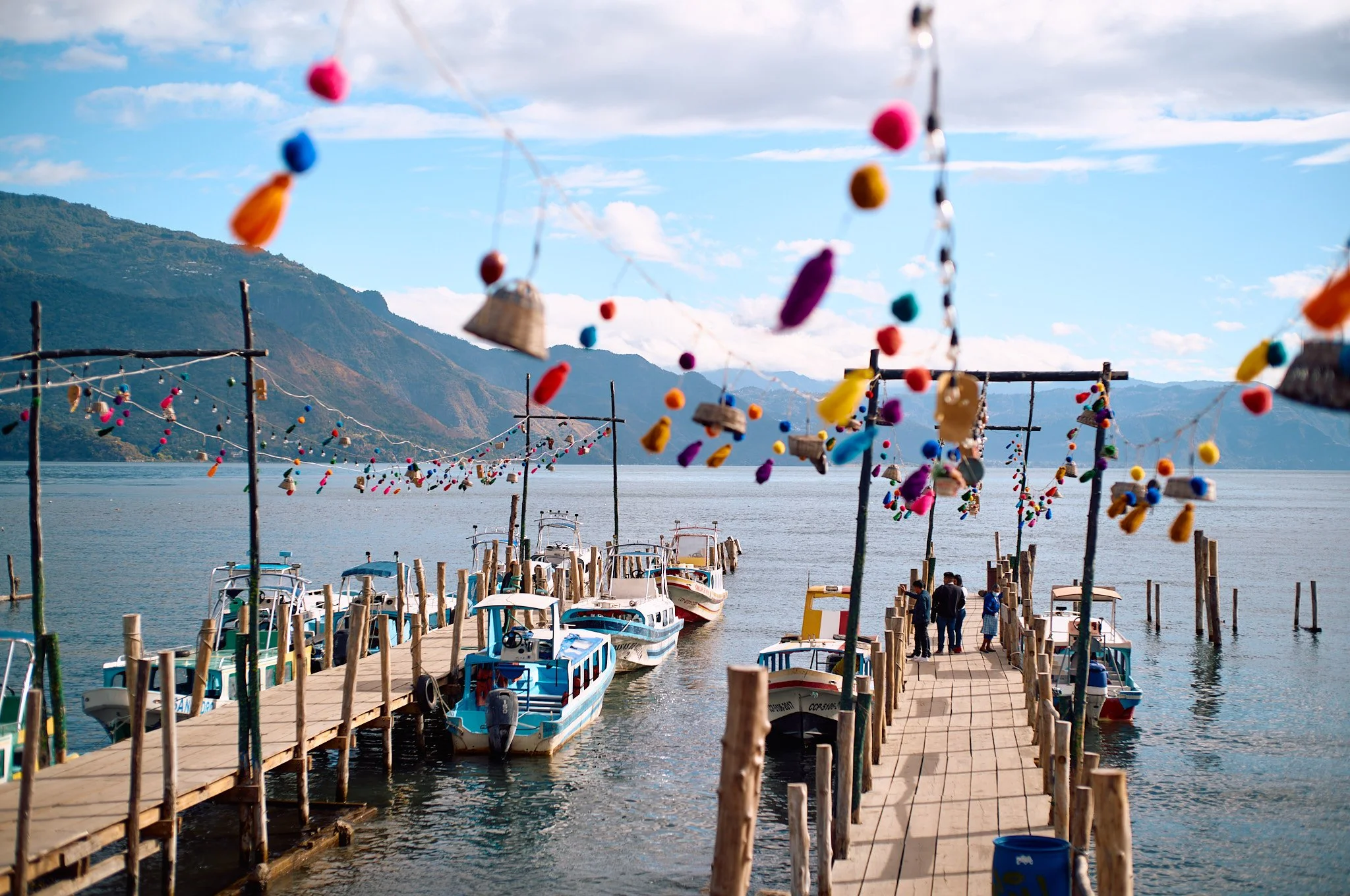 Boats docked at a wooden pier on a lake, decorated with colorful hanging ornaments and bottles, with mountains in the background under a partly cloudy sky.