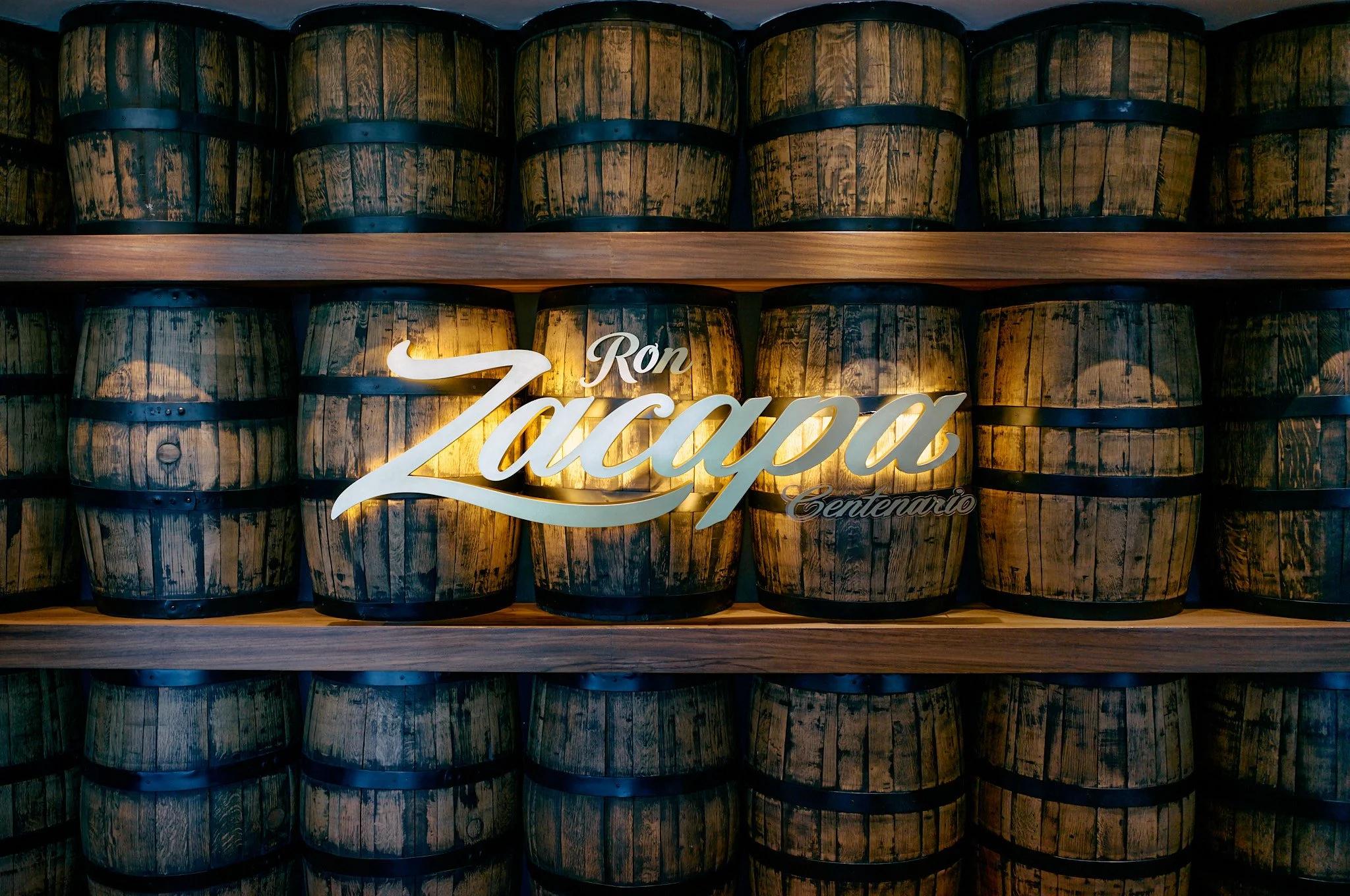 Wooden barrels stacked on shelves with a neon sign in front that reads 'Ron Zaccapa Centenario'.