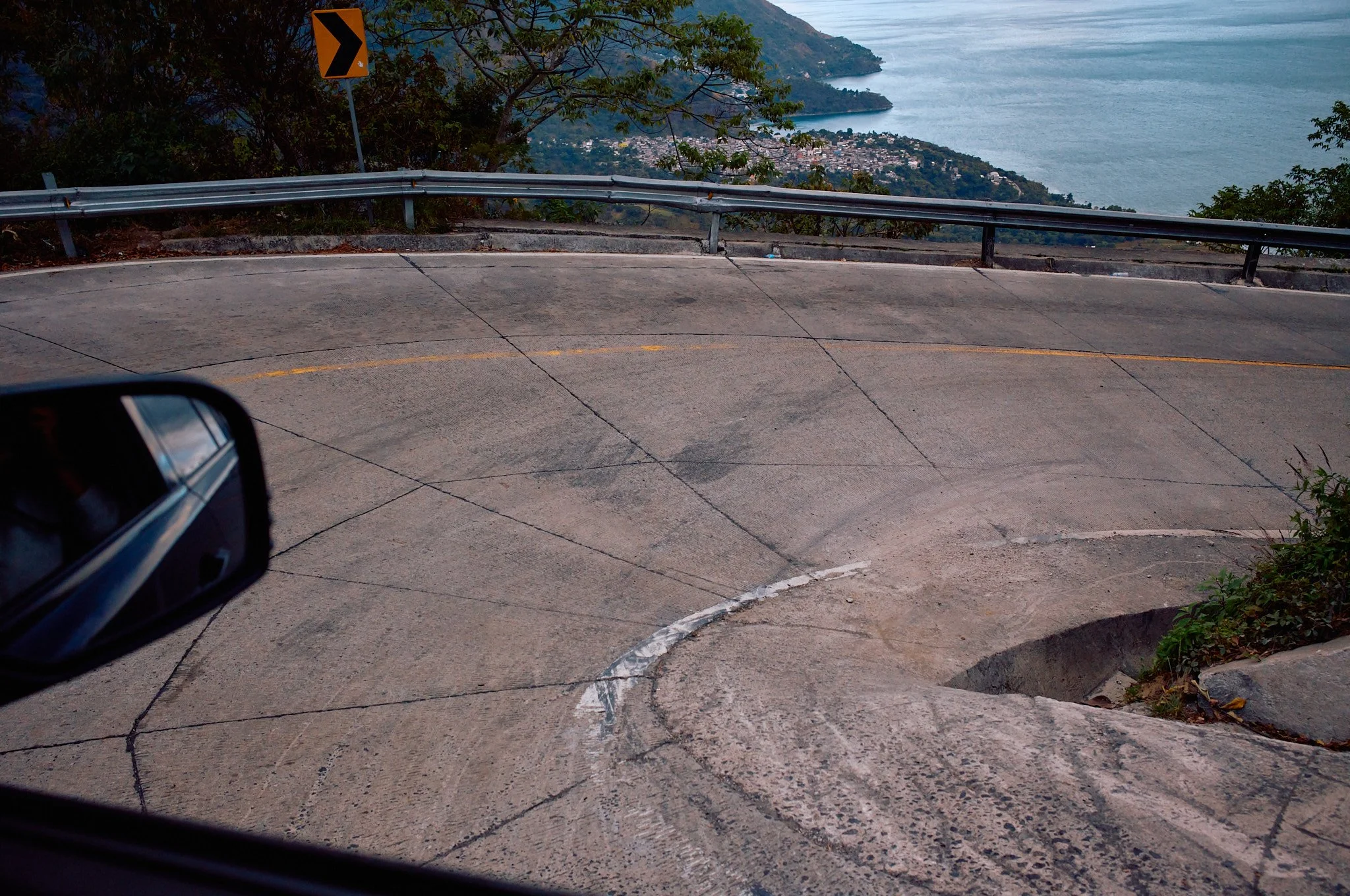 A winding mountain road with a sharp turn, guardrail, and a view of a large body of water and hills in the background, seen from inside a car.