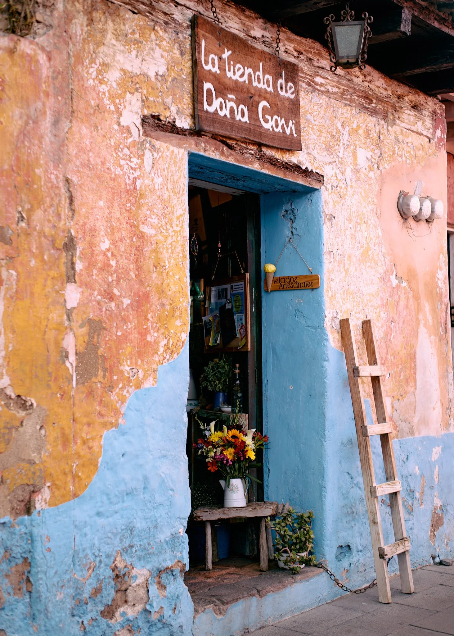 Small shop with a weathered turquoise and pink wall, a wooden sign that reads 'La tienda de Dña Gavi', a flower arrangement on a small bench outside, and a wooden ladder leaning against the wall.