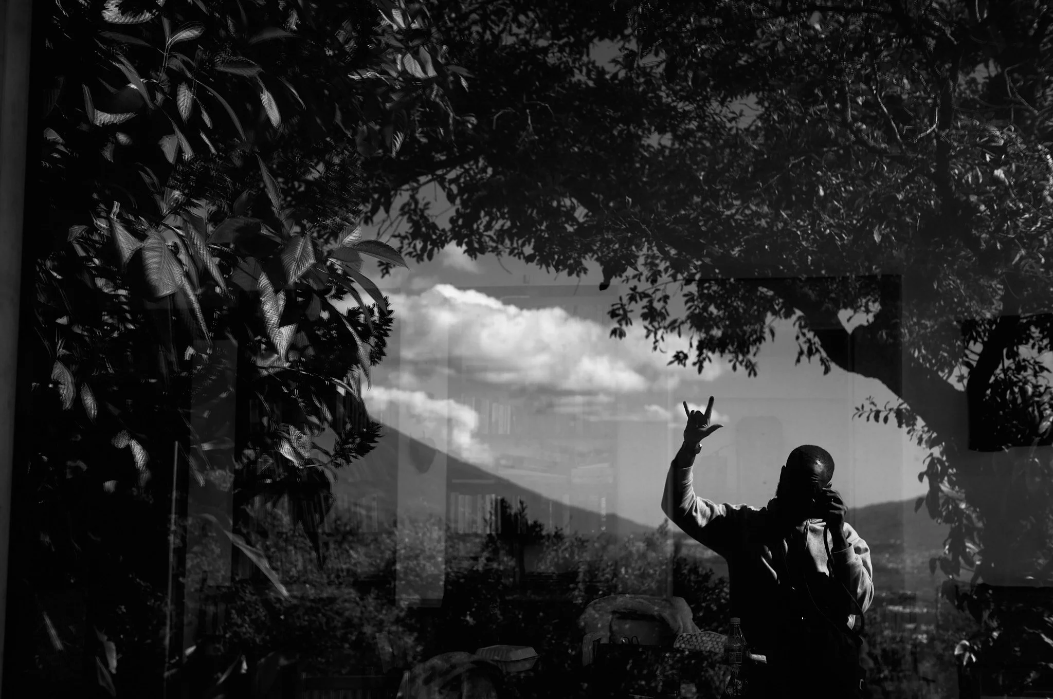 Silhouette of a person taking a photo with a smartphone, making a hand gesture, seen through a window, with trees and mountains in the background.
