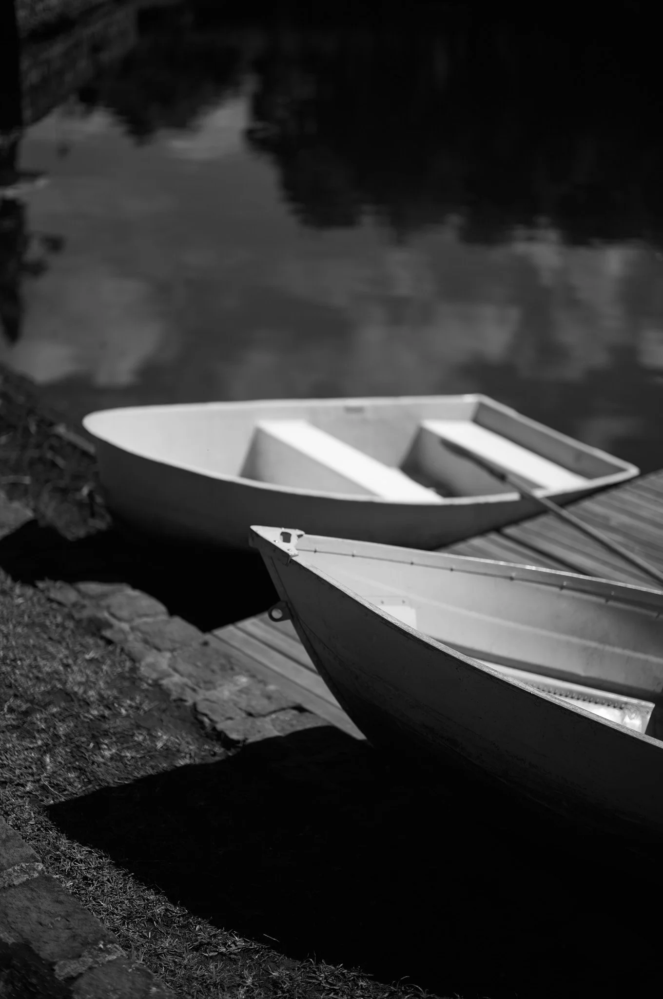 Two rowboats docked beside a stone embankment on a calm river, with reflections of trees and clouds in the water.