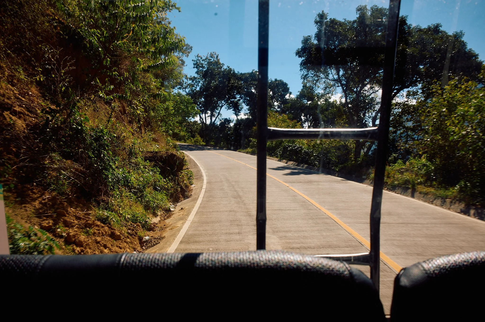 View of a winding mountain road with trees on both sides, seen from the back of a vehicle with a metal frame and black seats.
