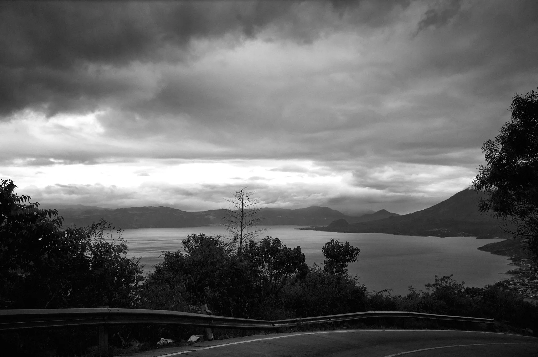 A black and white photo of a lake surrounded by mountains and trees, with a cloudy sky overhead.
