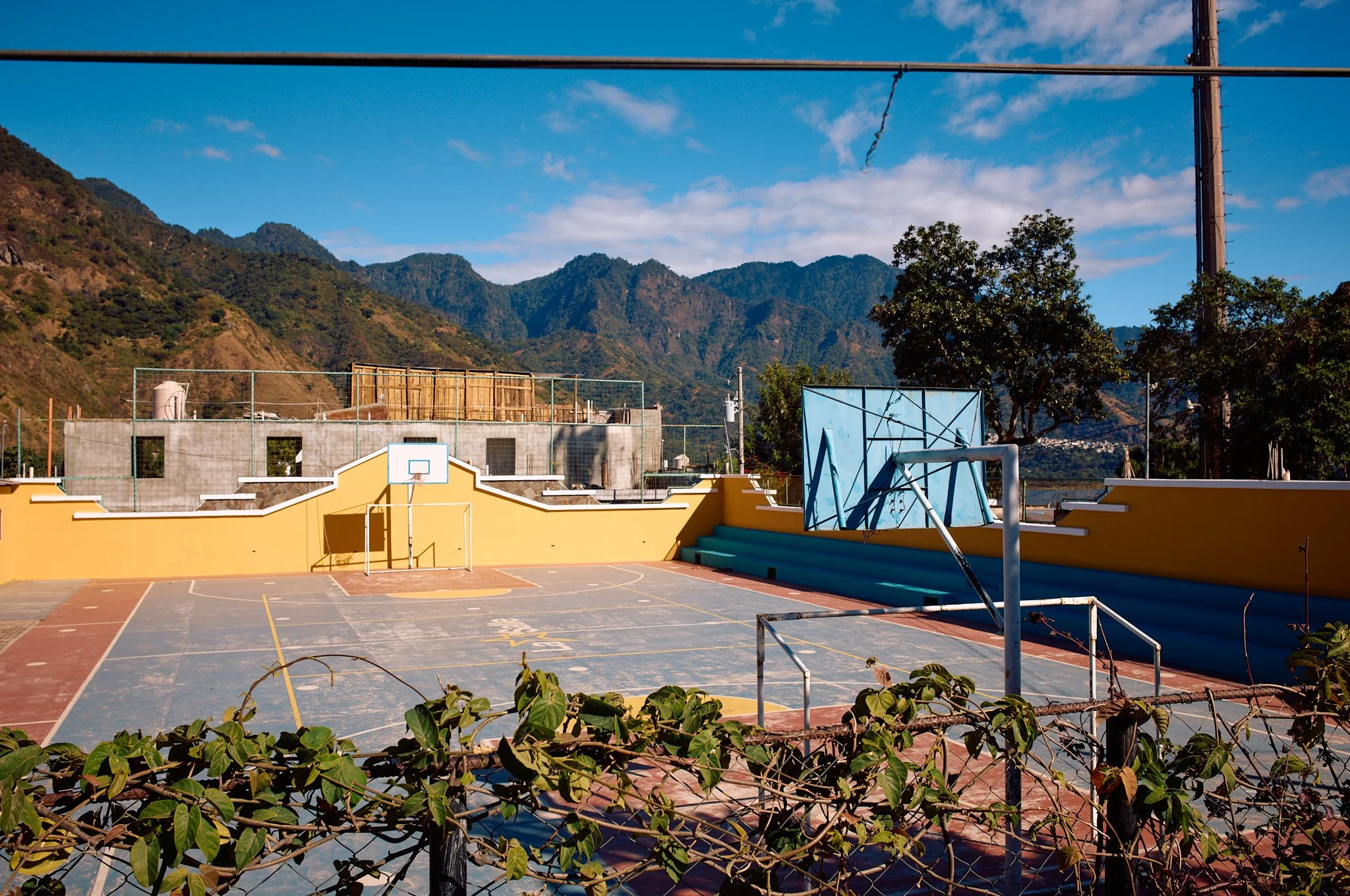 An outdoor basketball court with yellow walls, surrounded by a mountain landscape, with trees and blue sky in the background.