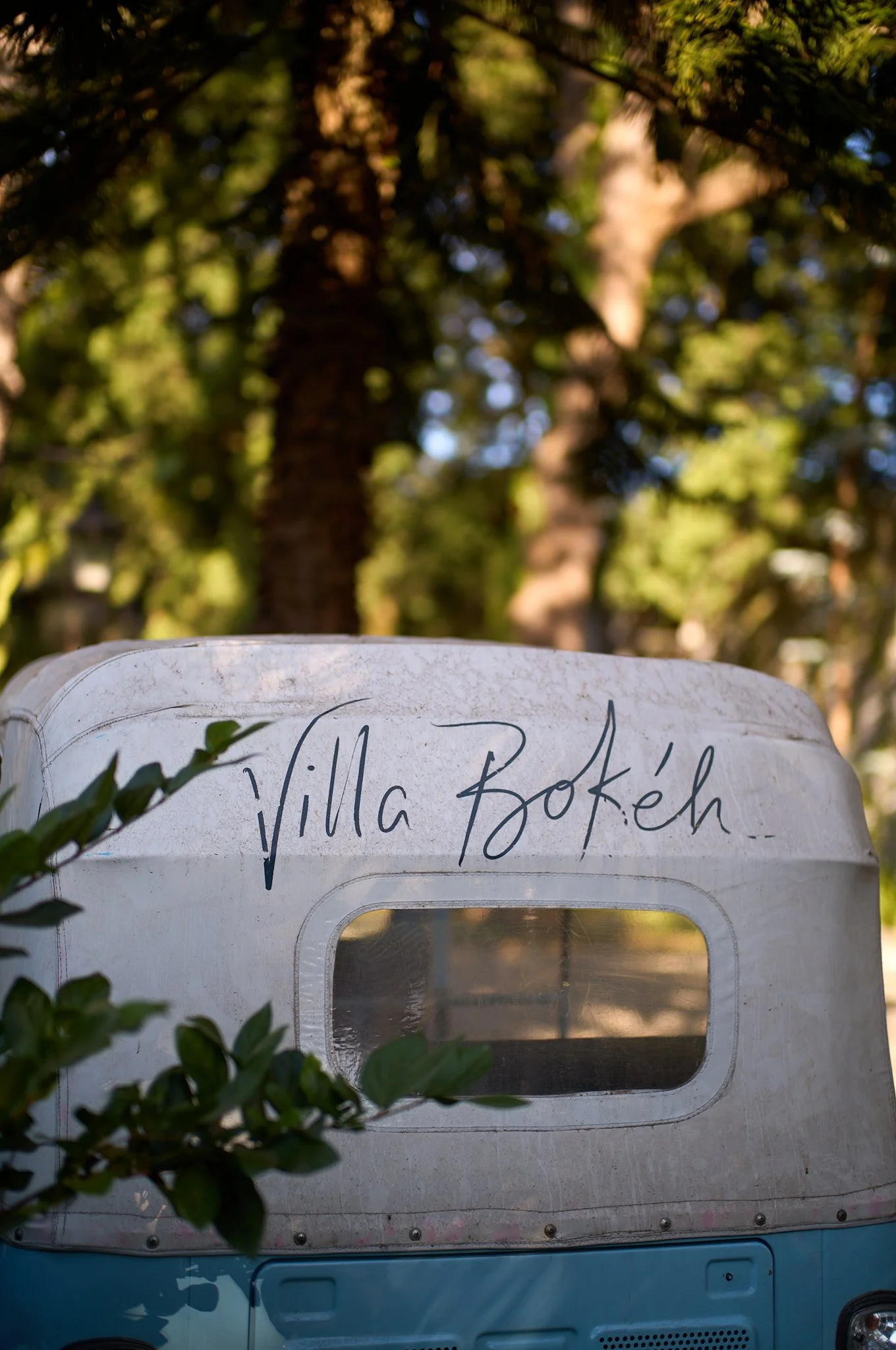 A vintage blue and white camper van with a window, parked among trees, with the handwritten text 'Villa Bokéh' on it.