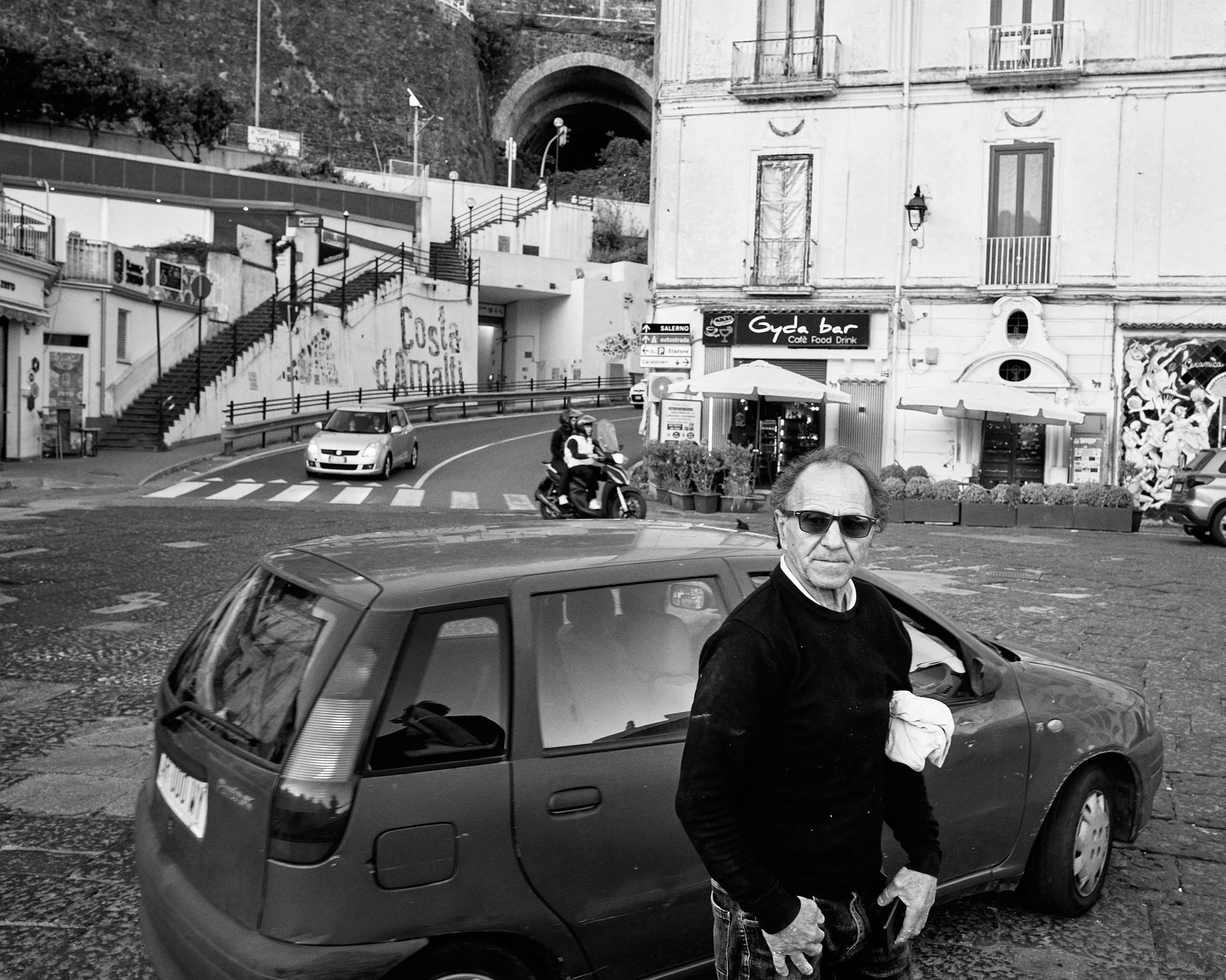 A man with sunglasses and a black sweater standing next to a small hatchback car on a cobblestone street, with buildings and people riding a scooter in the background.