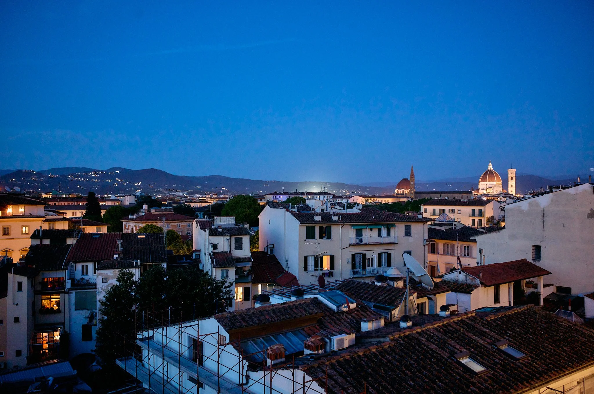 Nighttime cityscape of Florence, Italy, with illuminated historic buildings and the dome of the Florence Cathedral visible against a blue sky.