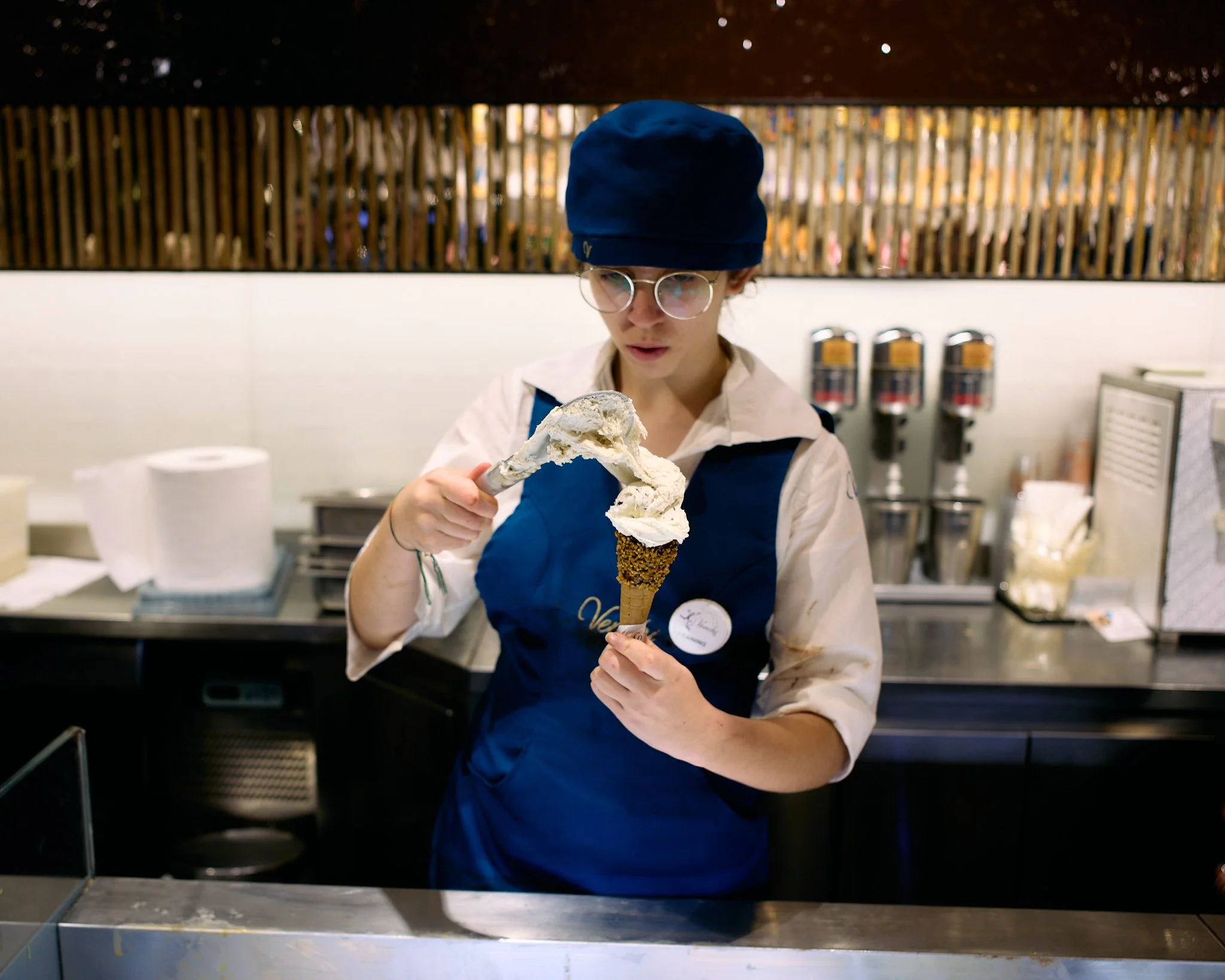 A woman wearing a blue apron, glasses, and a dark blue hat is holding an ice cream cone with vanilla ice cream in a store kitchen.