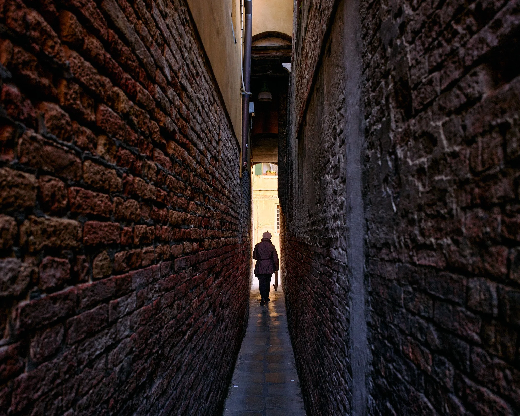 A narrow alleyway with brick walls on both sides and a person walking away from the camera toward a brightly lit area at the end of the alley.