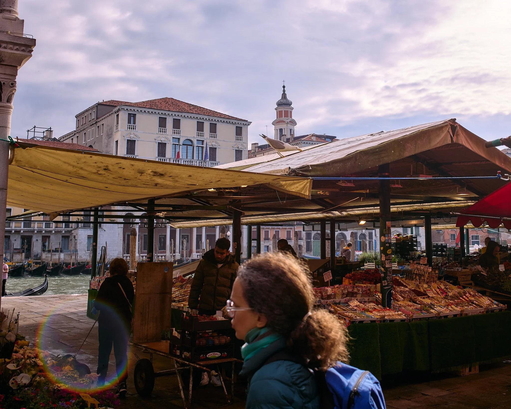 Venetian marketplace with people shopping, colorful produce and flowers, canal with gondolas, historic buildings, and a church tower in Venice, Italy.