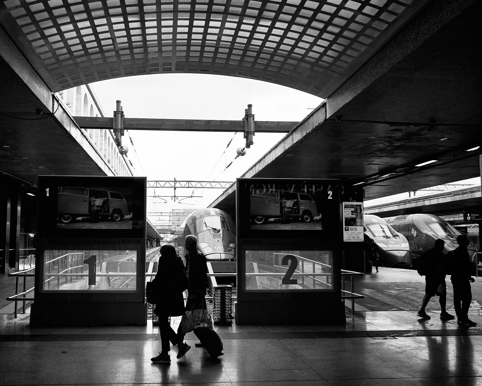 Black and white photo of a train station platform with high-speed trains in the background. Two people are walking with luggage in the foreground, silhouette against the bright background. There are signs labeled '1' and '2' for platforms, and digita