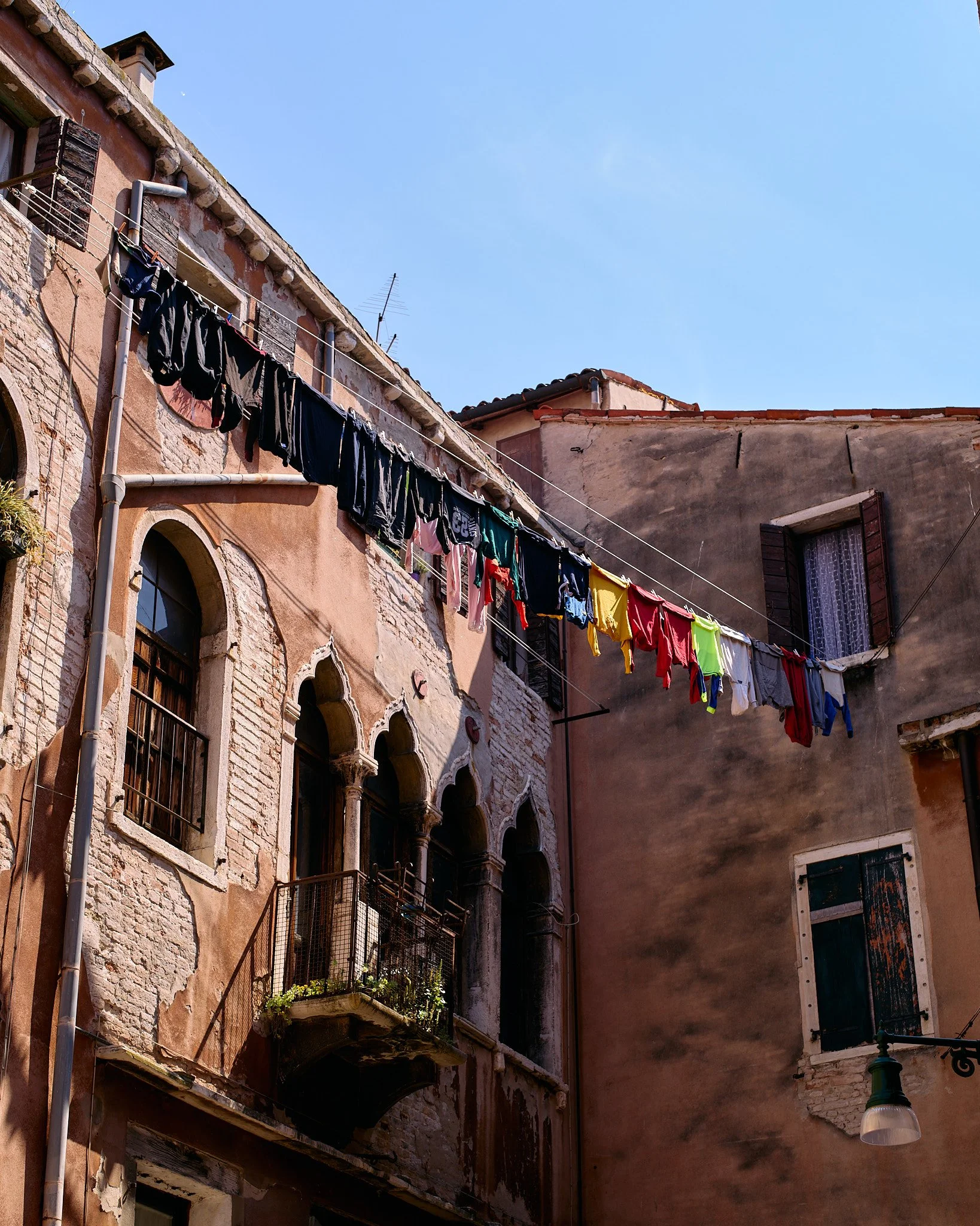 Clothes hanging on a line between old, weathered buildings with arched windows and balconies on a sunny day.