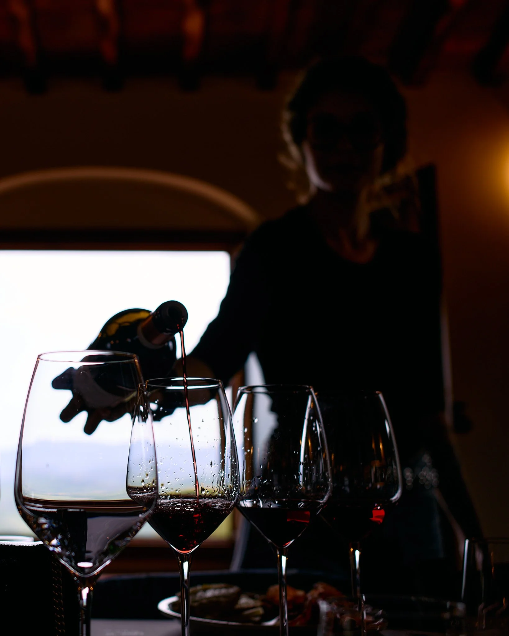 A person pouring red wine into glasses with a window in the background, creating a silhouette effect.