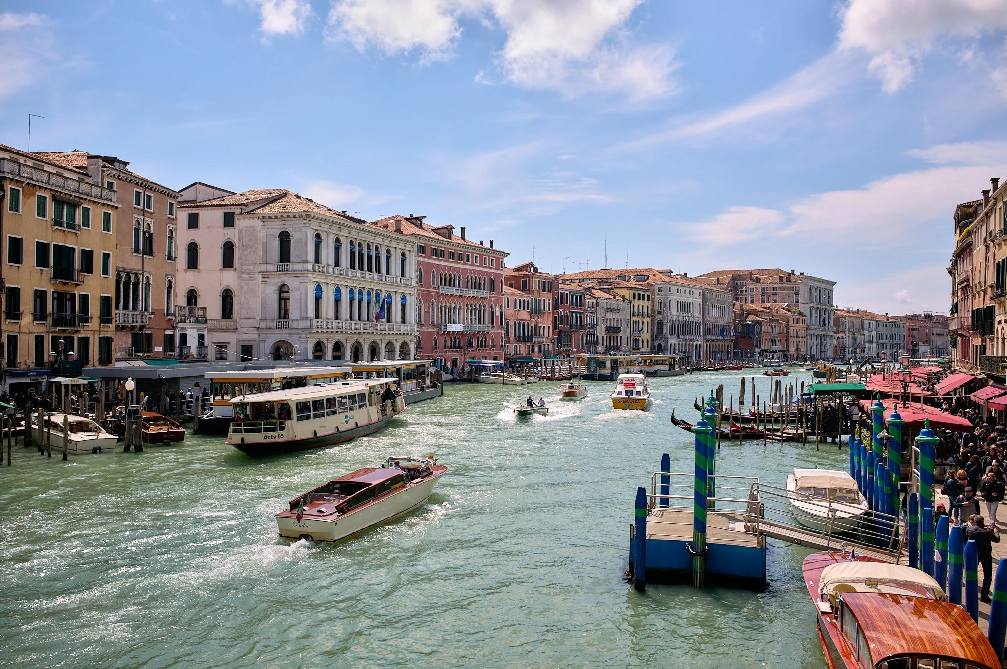 Venetian canal scene with boats, water taxis, colorful historic buildings lining the canal, and a clear blue sky.
