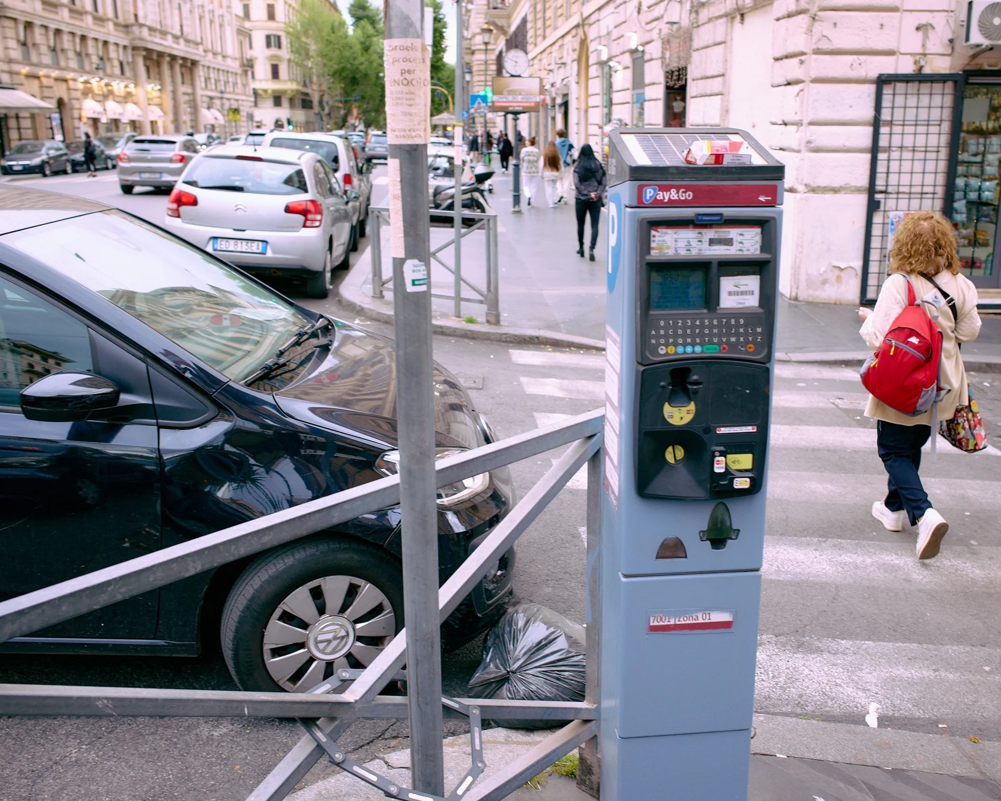 City street scene with parking meter, parked black car, sidewalk, people walking, and buildings in the background.
