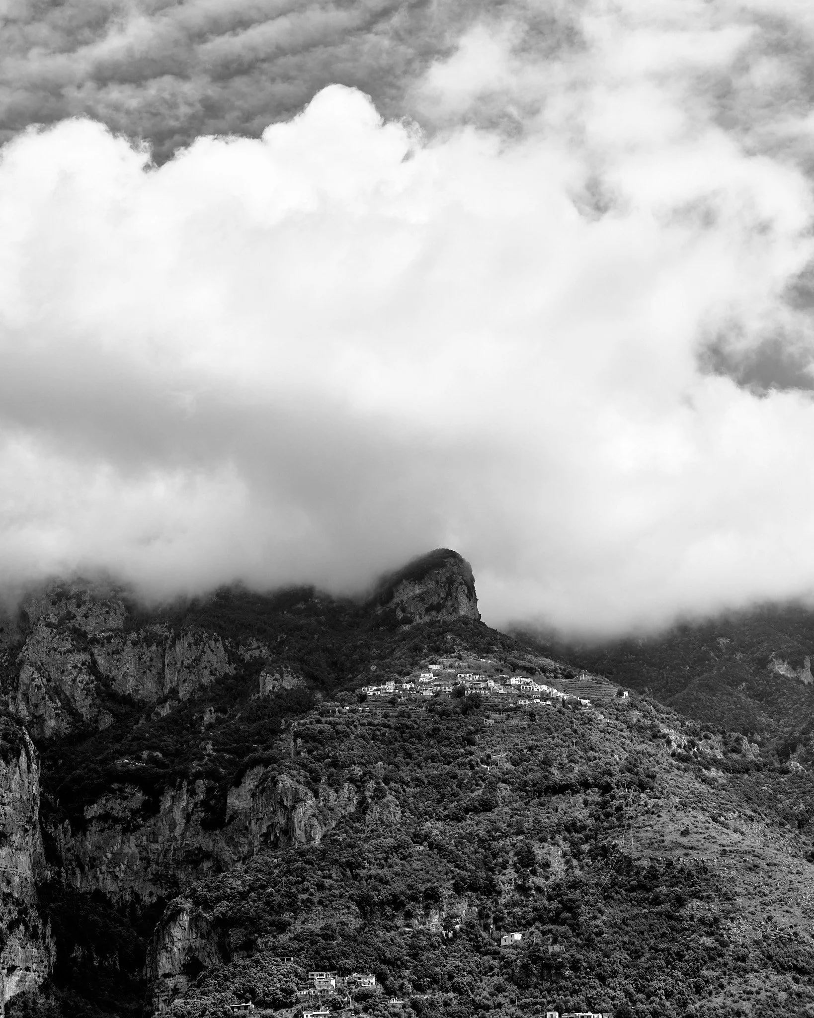 Black and white photograph of a mountain landscape in almafi with clouds covering the peaks and scattered buildings on the hillside.