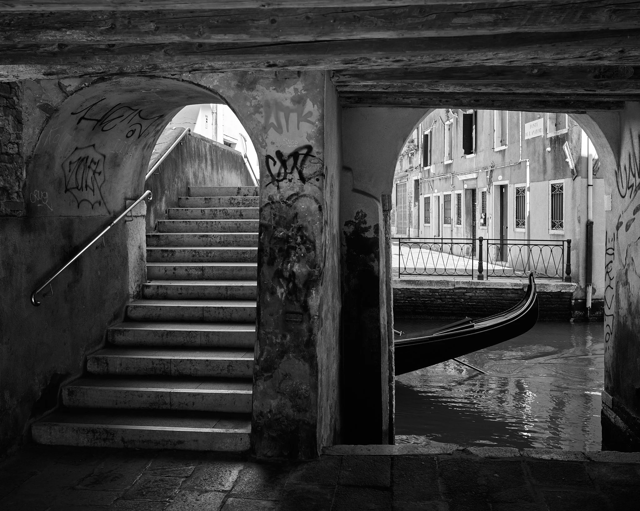 Black and white photo of a canal view in Venice, Italy, showing a staircase under a bridge with graffiti, a gondola in the water, and buildings along the canal with balconies and windows.