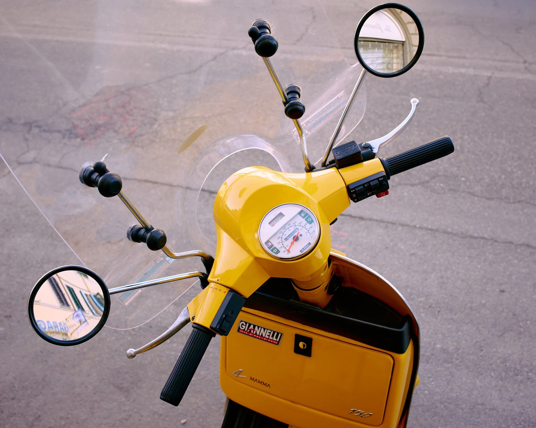 Top view of a yellow vintage scooter with rear and front mirrors, windshield, speedometer, and handlebar controls, parked on asphalt.