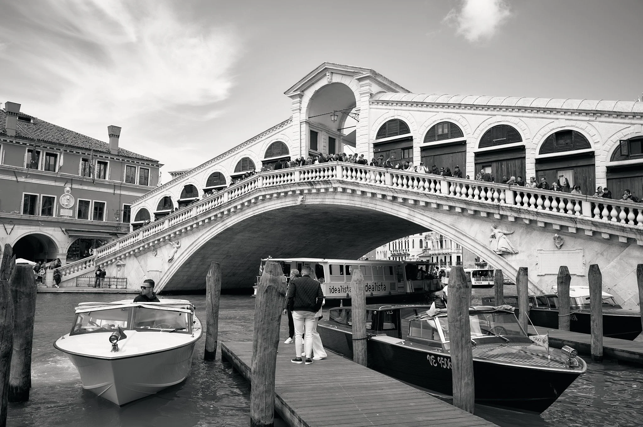 A black and white photo of the Rialto Bridge in Venice with boats docked below and people walking on the bridge.