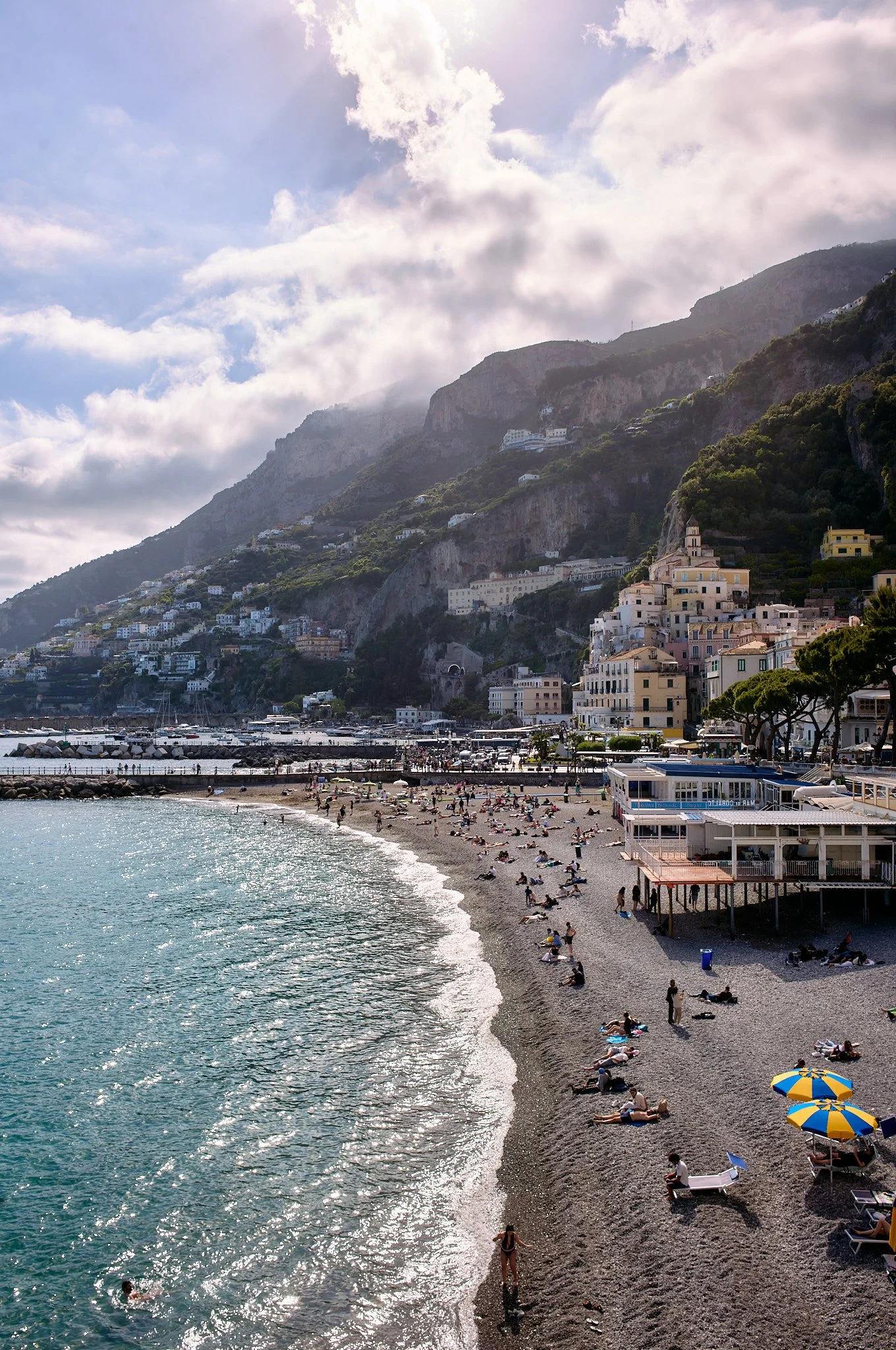 A scenic view of a pebble beach in Positano along the coastline of a hilly area, with buildings scattered on the hillside and a partly cloudy sky overhead.