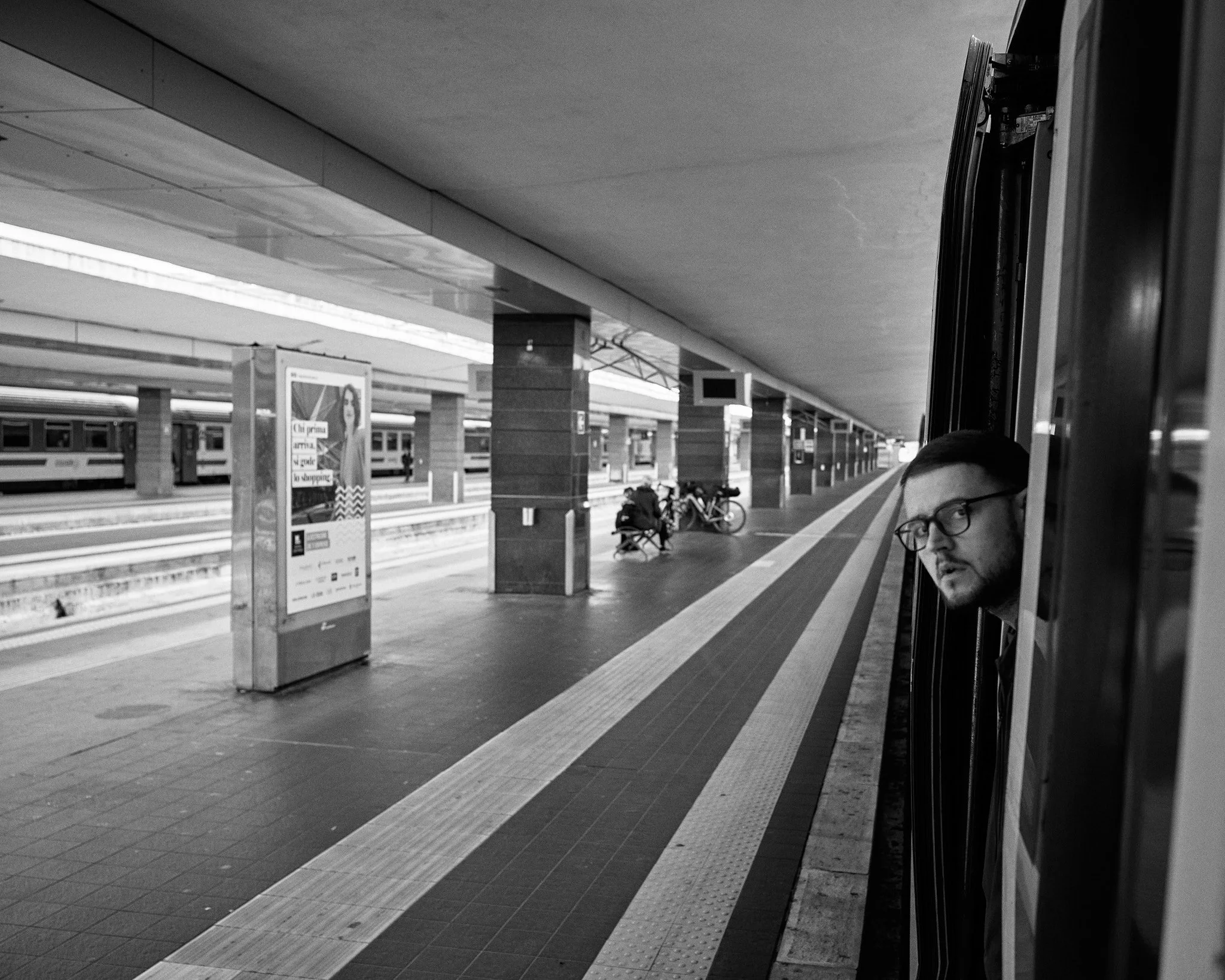 A man with glasses looking out of a moving train at a train station platform in black and white.