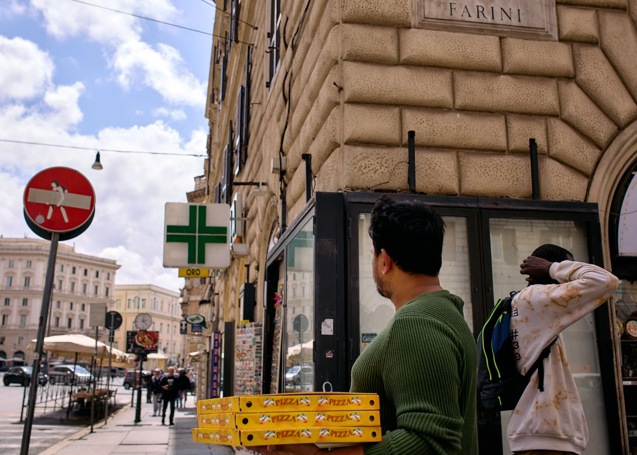 Two men are on a city sidewalk, one in a green sweater holding stacked pizza boxes, the other adjusting his hair with a backpack. Behind them, a building has a sign labeled "FARINI" and a green cross sign of a pharmacy. There is also a no entry traff