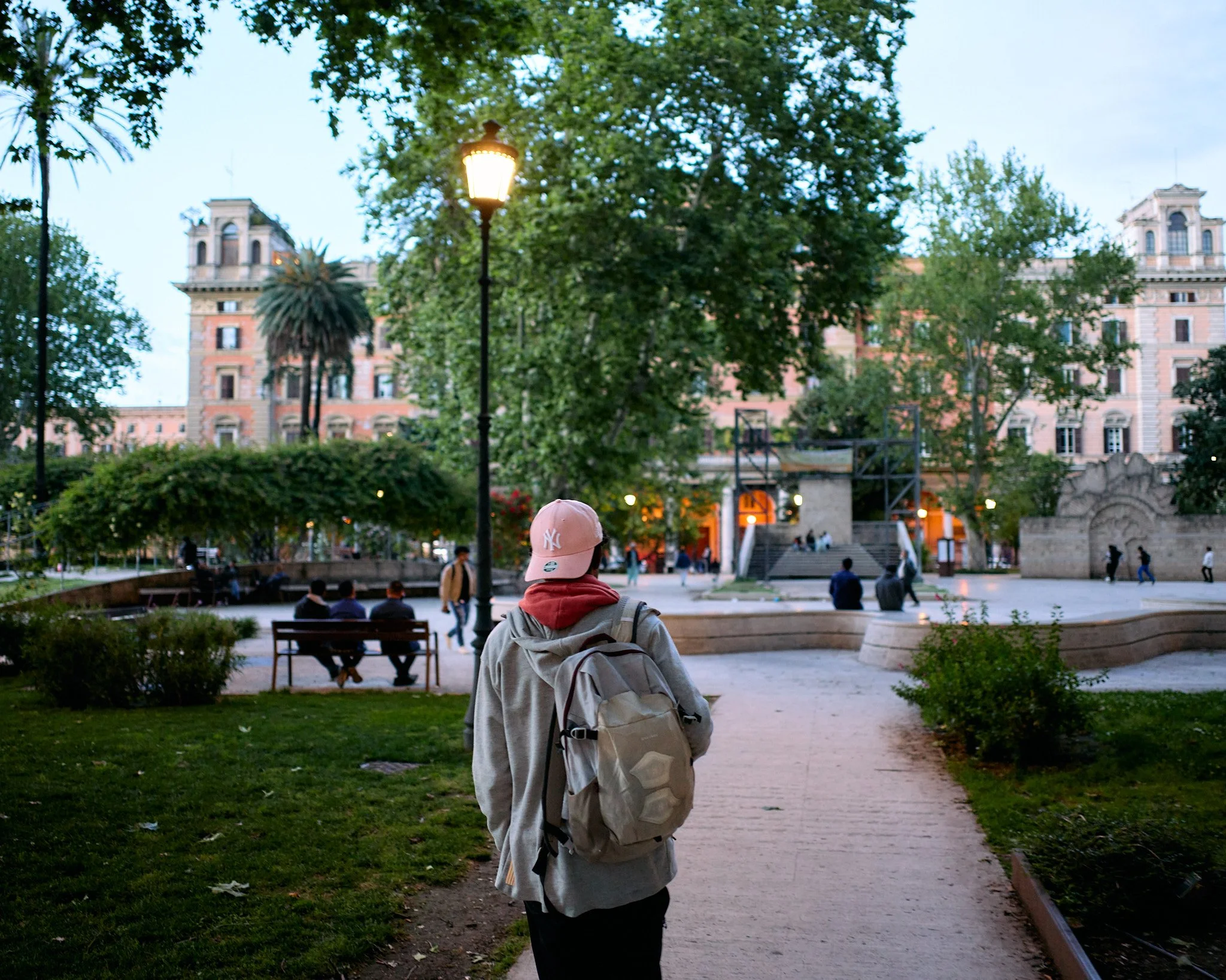 A person with a backpack walking on a park path during evening. Park benches and people are visible, with historic buildings in the background.