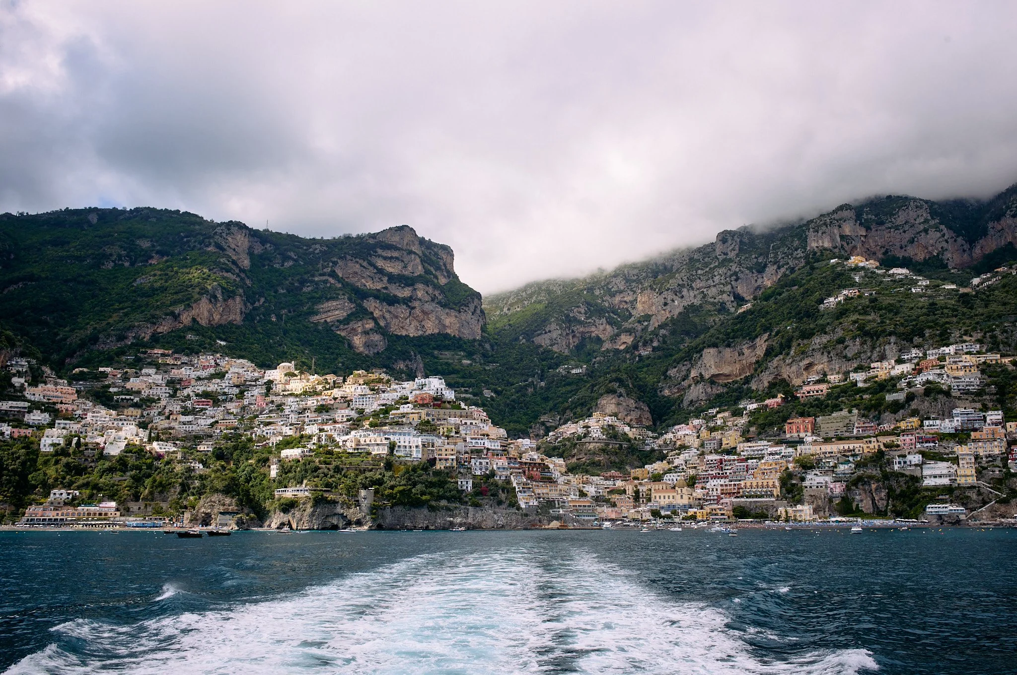 View of a coastal town with colorful buildings on a cliffside, overlooking the sea with boat wakes, and green mountains topped with clouds in the background.