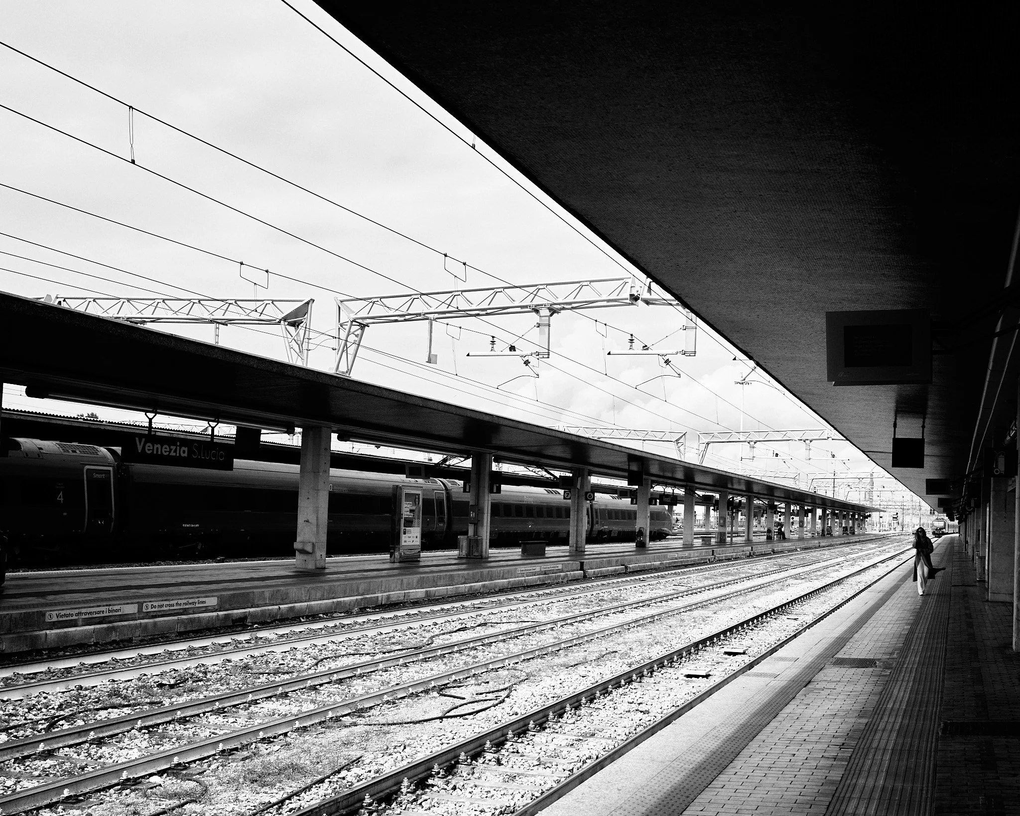 Black and white photo of an empty train station platform with a single person waiting. Multiple train tracks run parallel, with a train on the opposite side. Overhead signage reads "Venezia S. Lucia." Overhead electrical lines and station infrastruct
