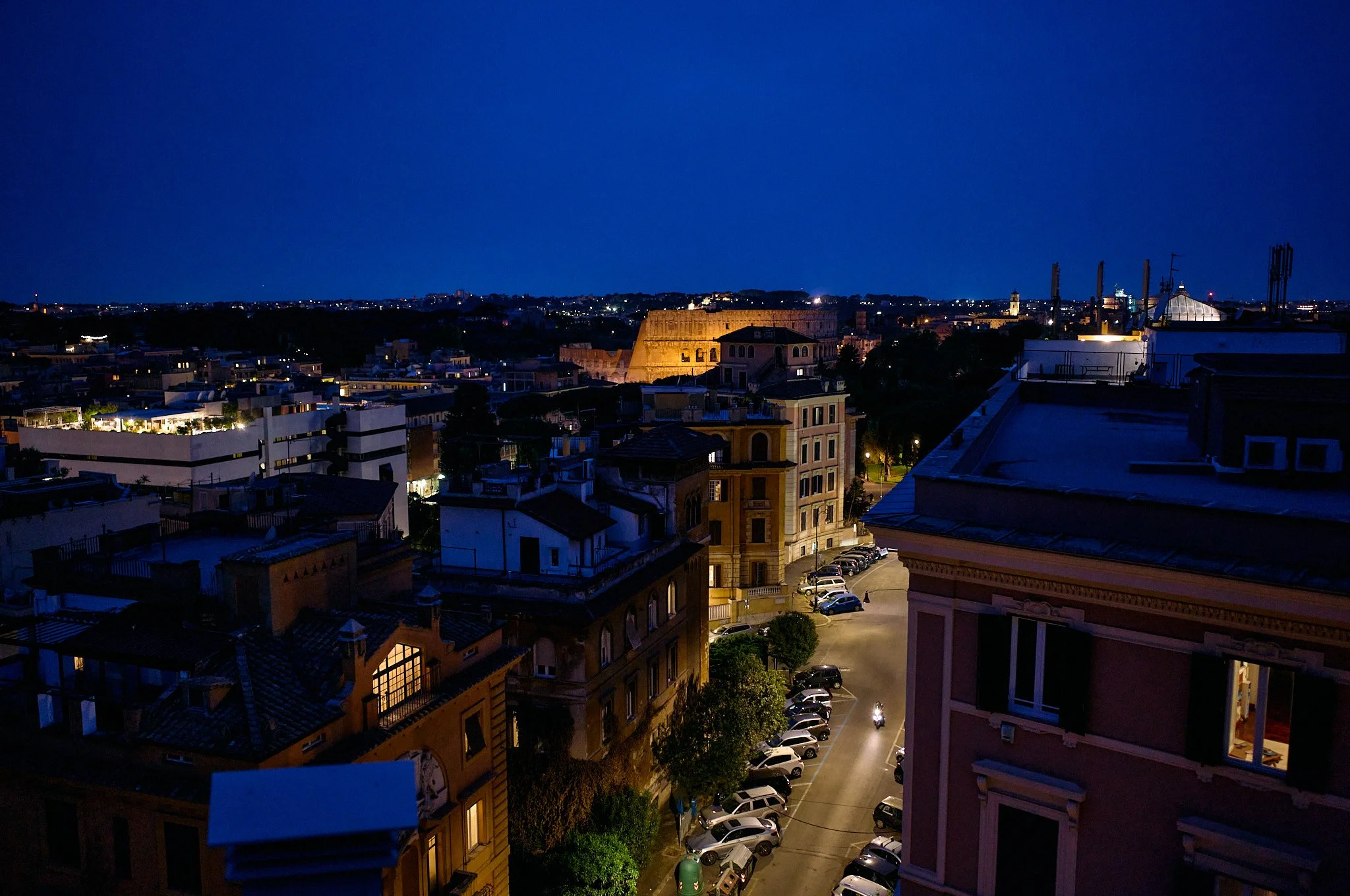 Nighttime cityscape view with buildings, roads, parked cars, and illuminated windows under a dark blue sky.