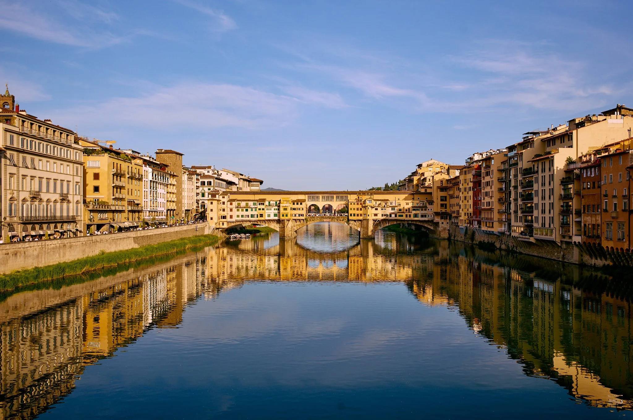 Photo of the historic Ponte Vecchio bridge over the Arno River in Florence, Italy, with colorful buildings lining the riverbanks and a clear blue sky above.