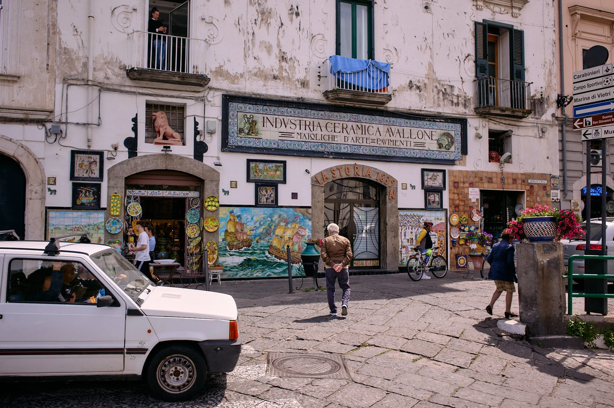 A street scene in Italy with a small ceramic shop called La Storia 1839, decorated with colorful artwork and plates. There are people walking, a man riding a bicycle, and an older man facing the shop. A parked white car is in the foreground, and buil