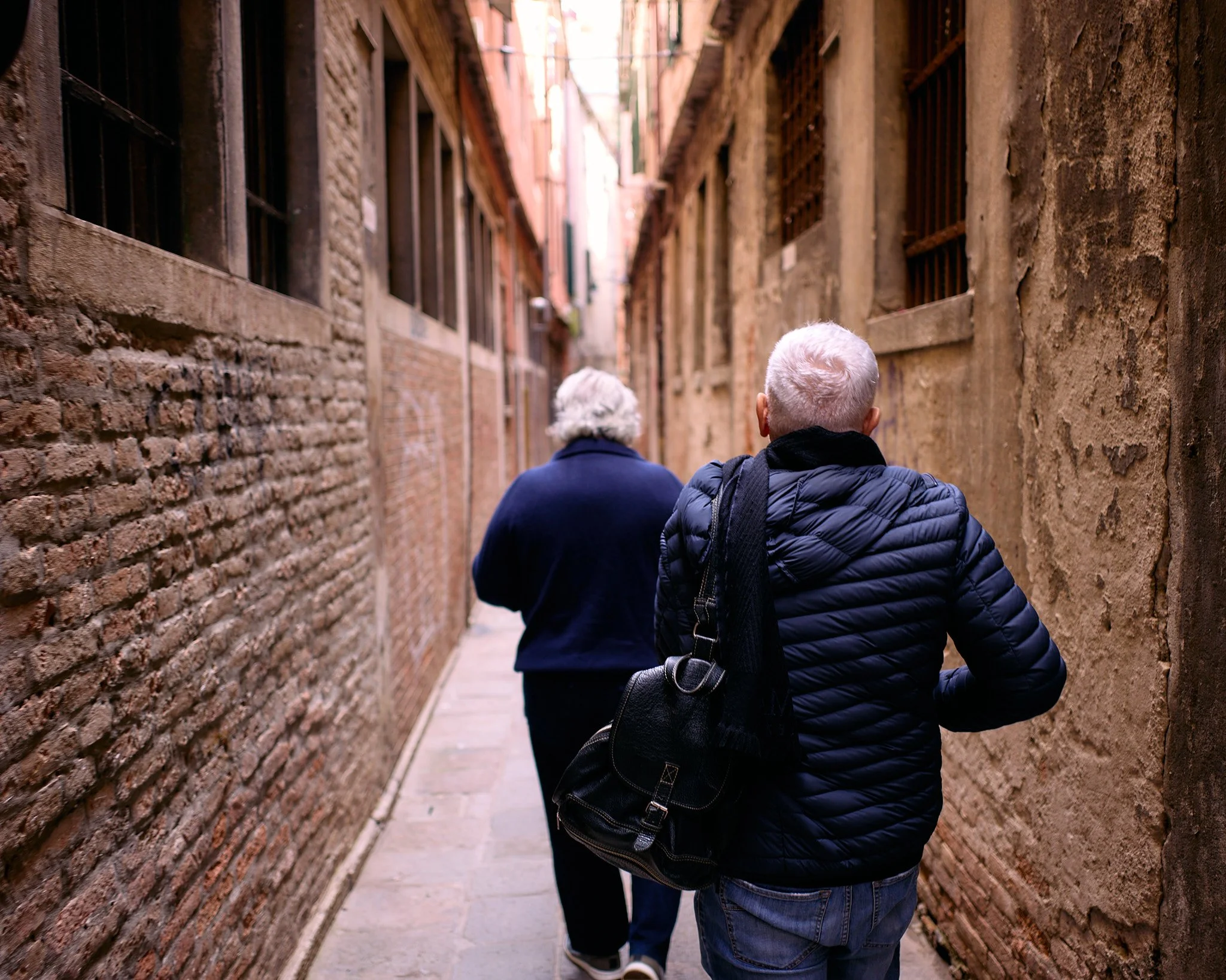 Two people walking through a narrow alleyway with brick and stucco walls, one with white hair and a black backpack, the other with curly white hair and a navy coat.