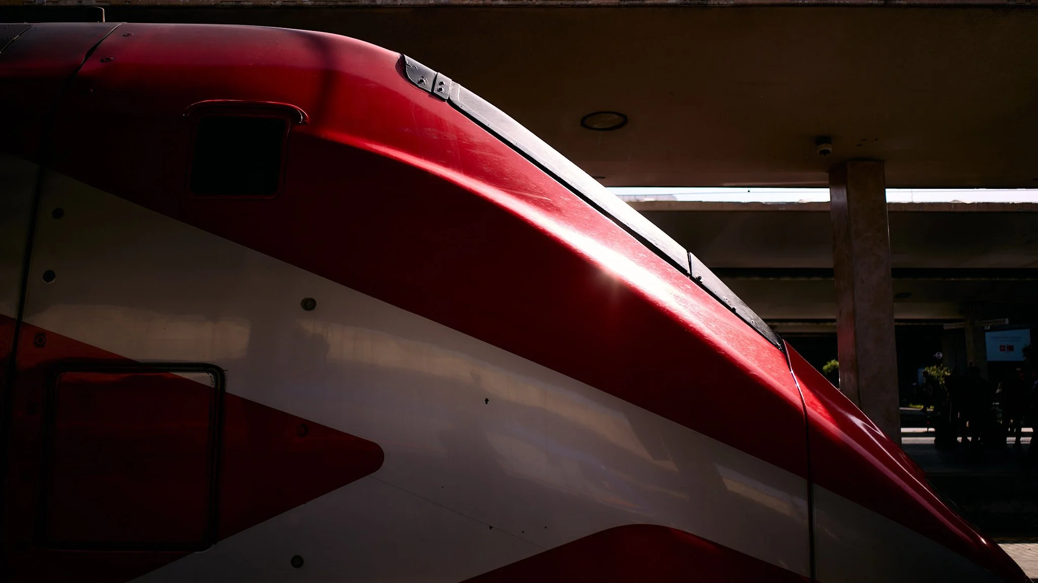 Close-up of a red and white race car with sunlight reflecting off its surface, under a covered structure.