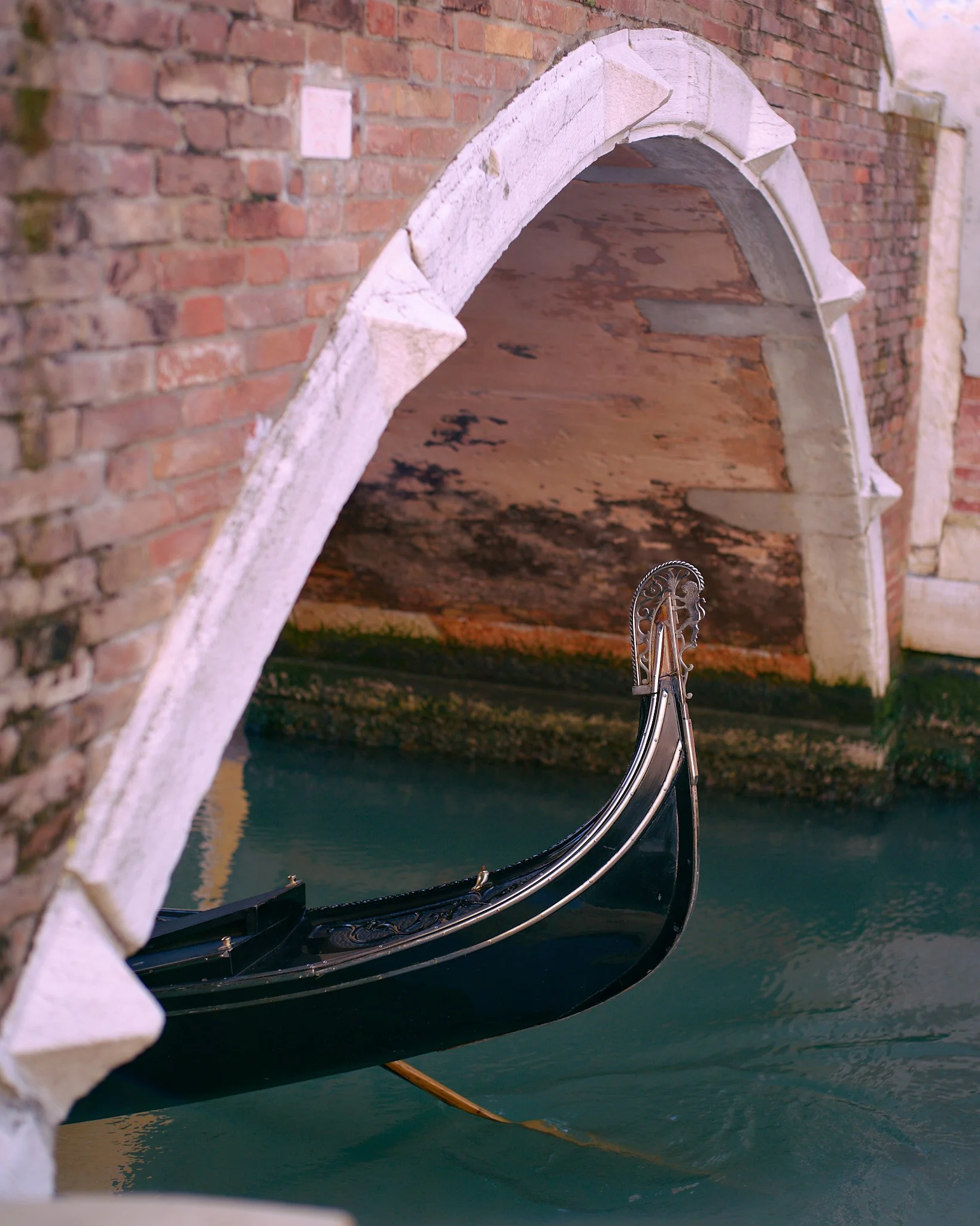 A Venetian canal with a black gondola moored under a brick and white stone bridge, with water reflecting the surroundings.