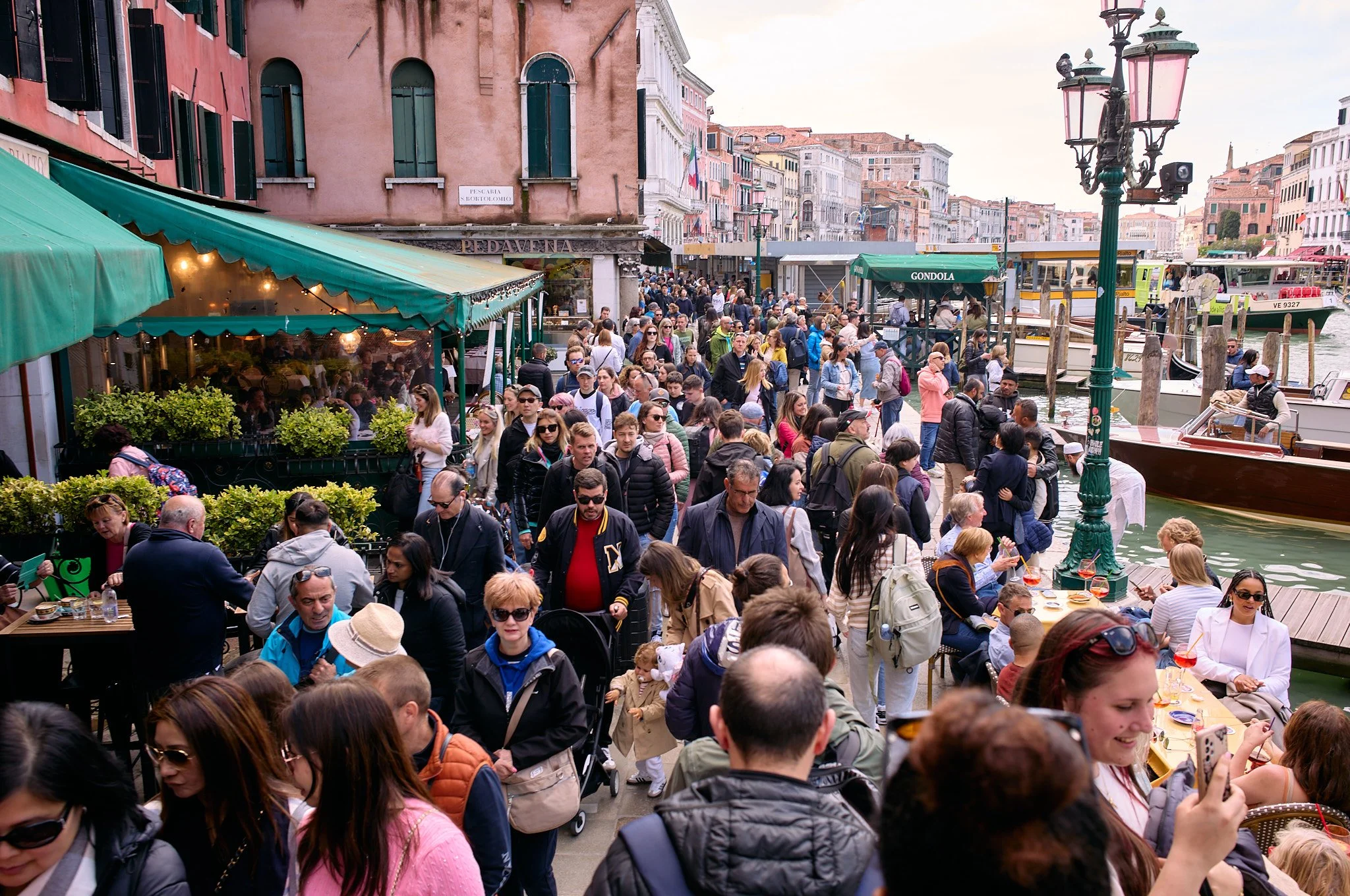 A crowded outdoor scene along a waterfront with many people walking, sitting, and enjoying food at cafes. There are boats on the water, a green canopy, street lamps, and colorful historic buildings in the background.