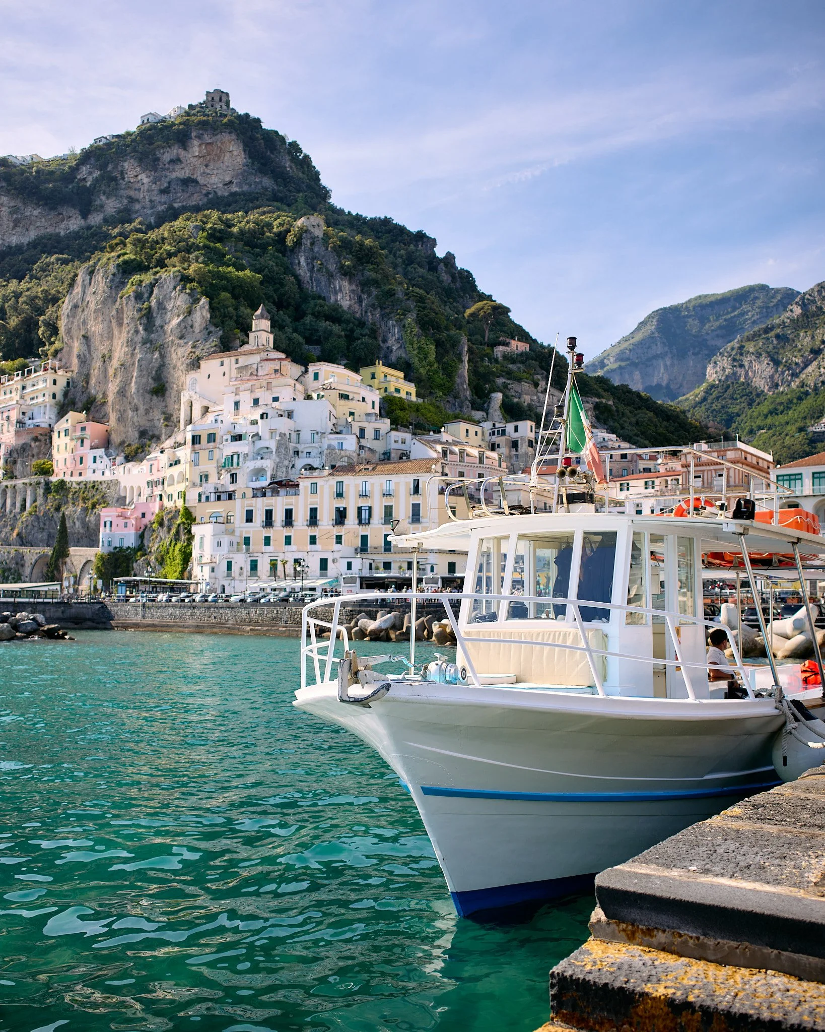 A small white boat docked at a harbor with colorful buildings on a hillside in the background. Mountains are visible behind the buildings, and the sky is partly cloudy.