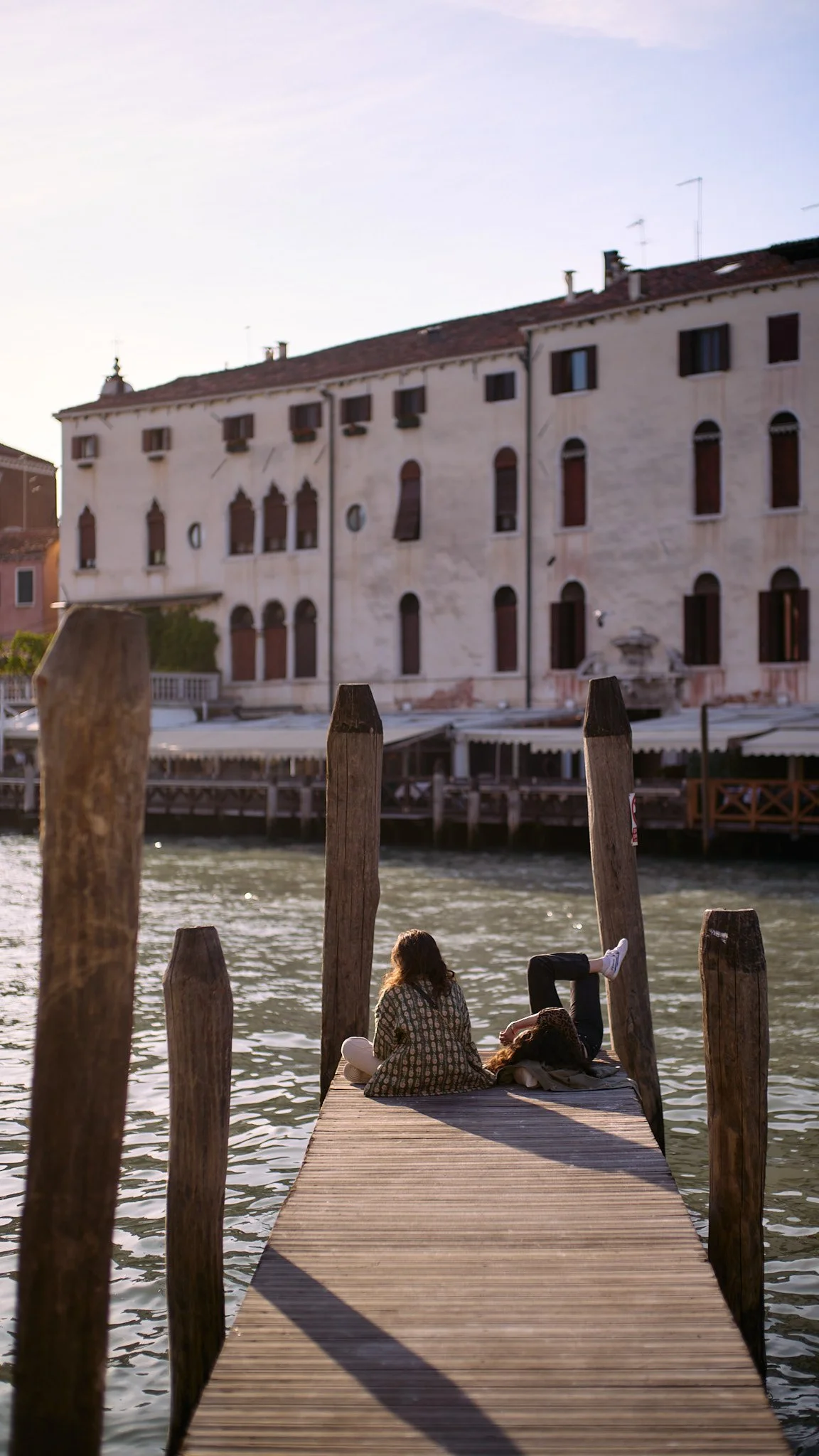 Two women sitting on a dock beside the water, with a large old building in the background.