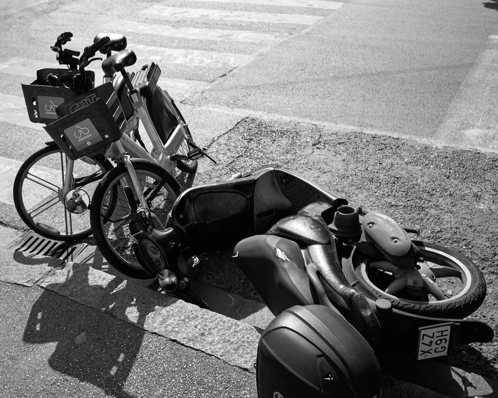 A fallen three-wheeled scooter with a bike nearby on the sidewalk, with bike rental racks in the background.