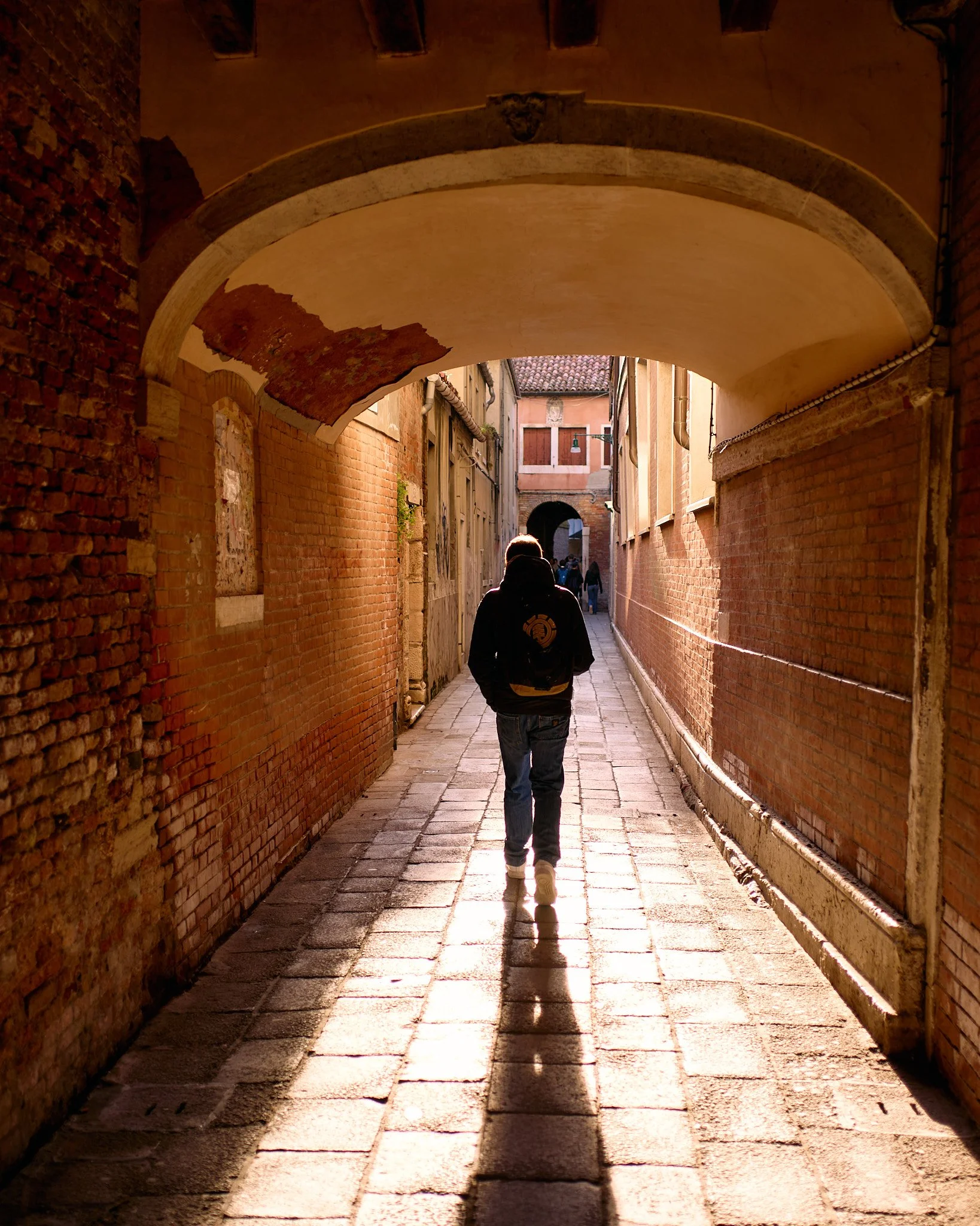 A person walking through a narrow, brick alleyway under an archway in an old European city, with sunlight casting long shadows.