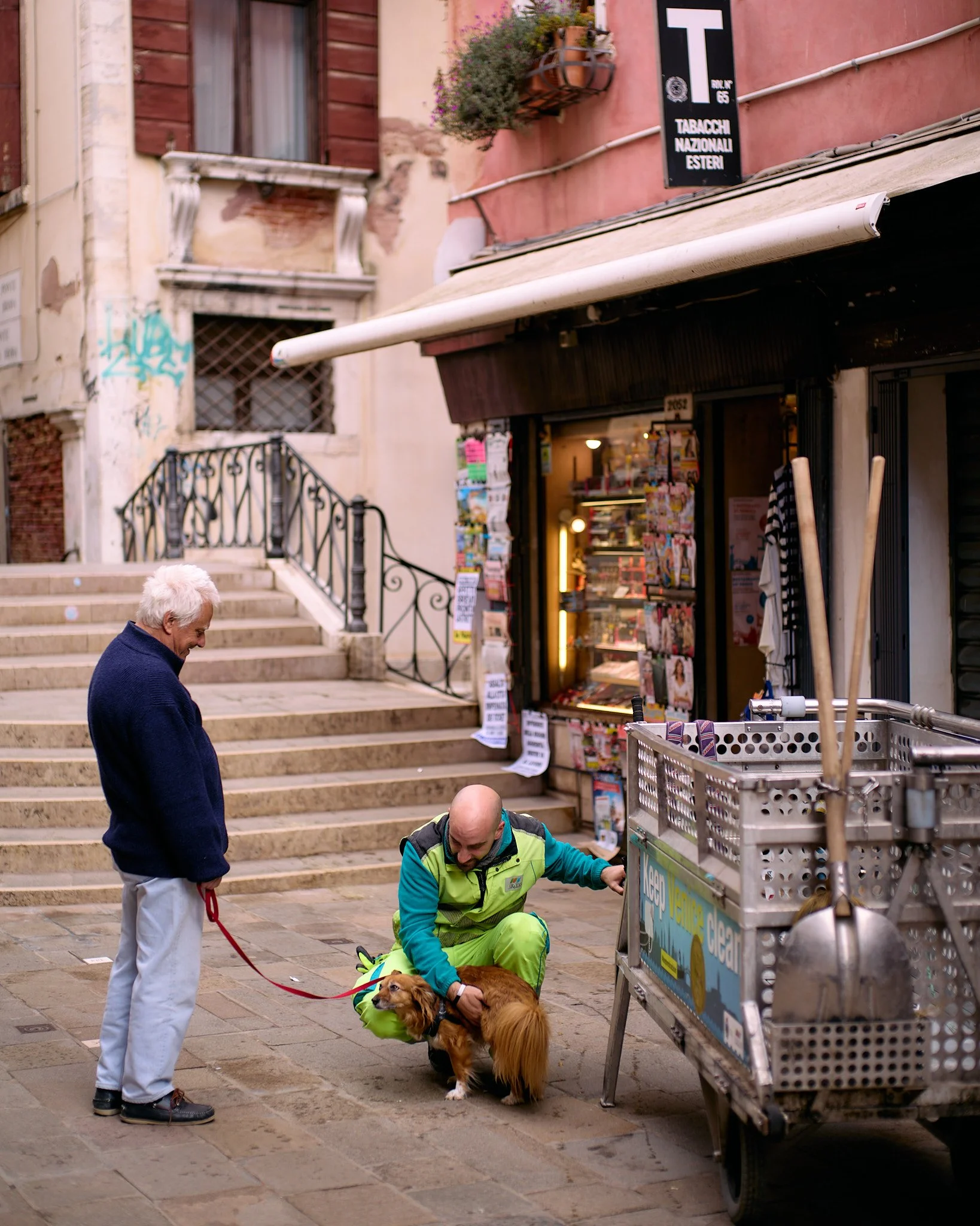 An elderly man holding a red leash, standing on cobblestone street; a man in a green jacket crouching down with a small brown dog near a newsstand; buildings with stairs and graffiti in the background.