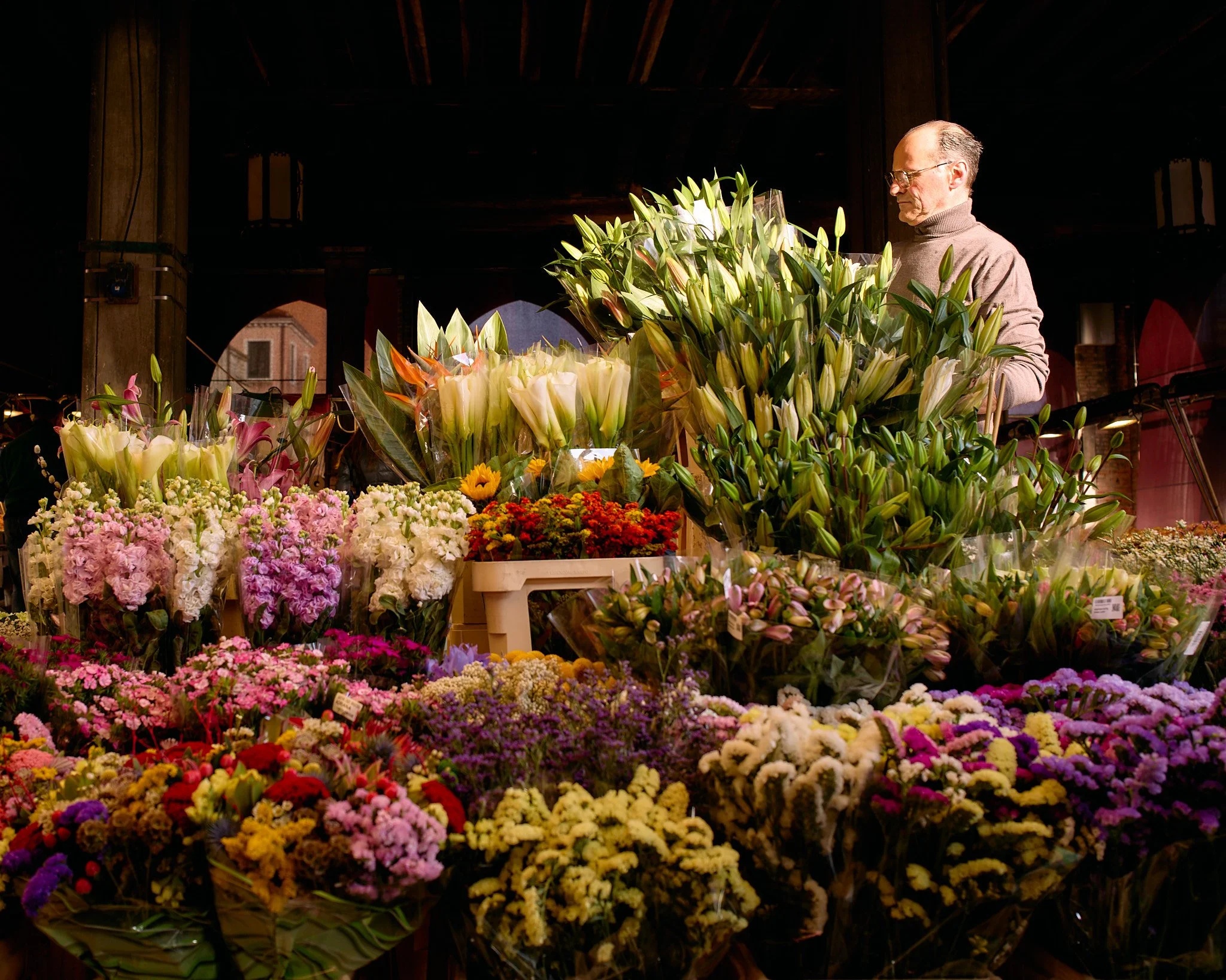 A man in glasses and a beige sweater arranging flowers in a flower shop with a variety of colorful flowers and lilies on display.