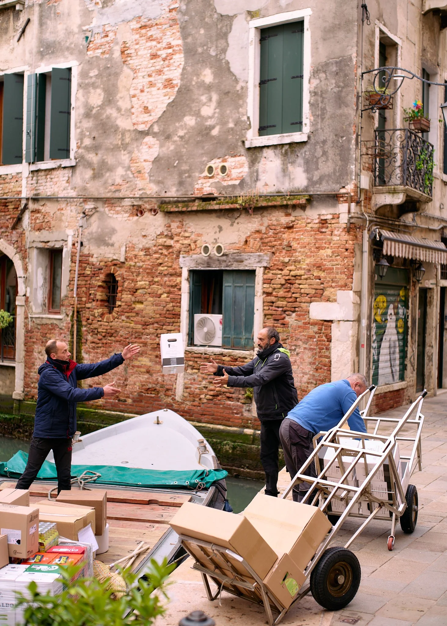 Three men are on a canal-side street in Venice, Italy, with a weathered brick building in the background. They are engaged in a conversation, with one man handing a white object to another. There are boxes and carts loaded with goods nearby.
