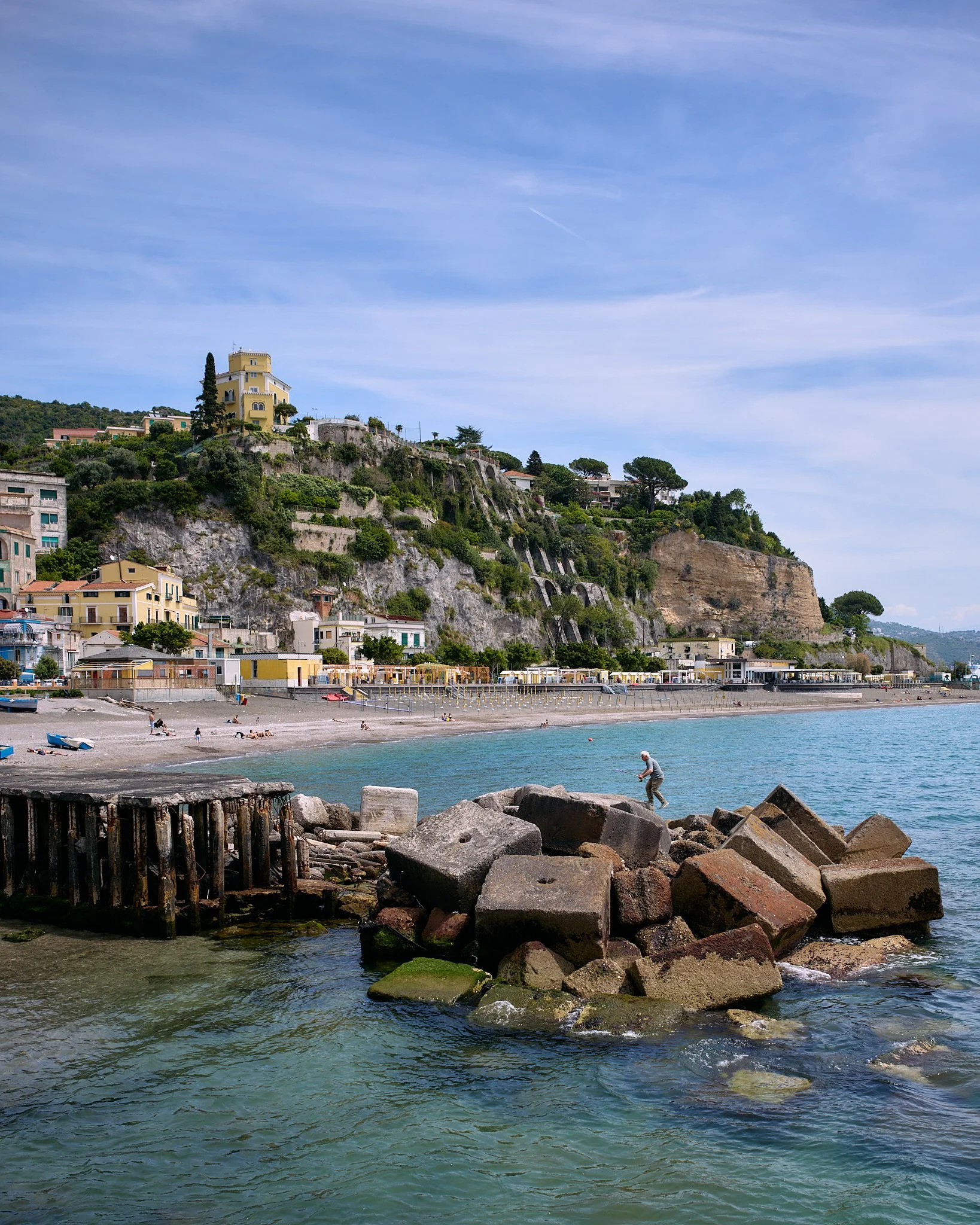 View of a seaside town with buildings on hills, a beach with people relaxing, and a man fishing on rocks in the water, under a partly cloudy sky.