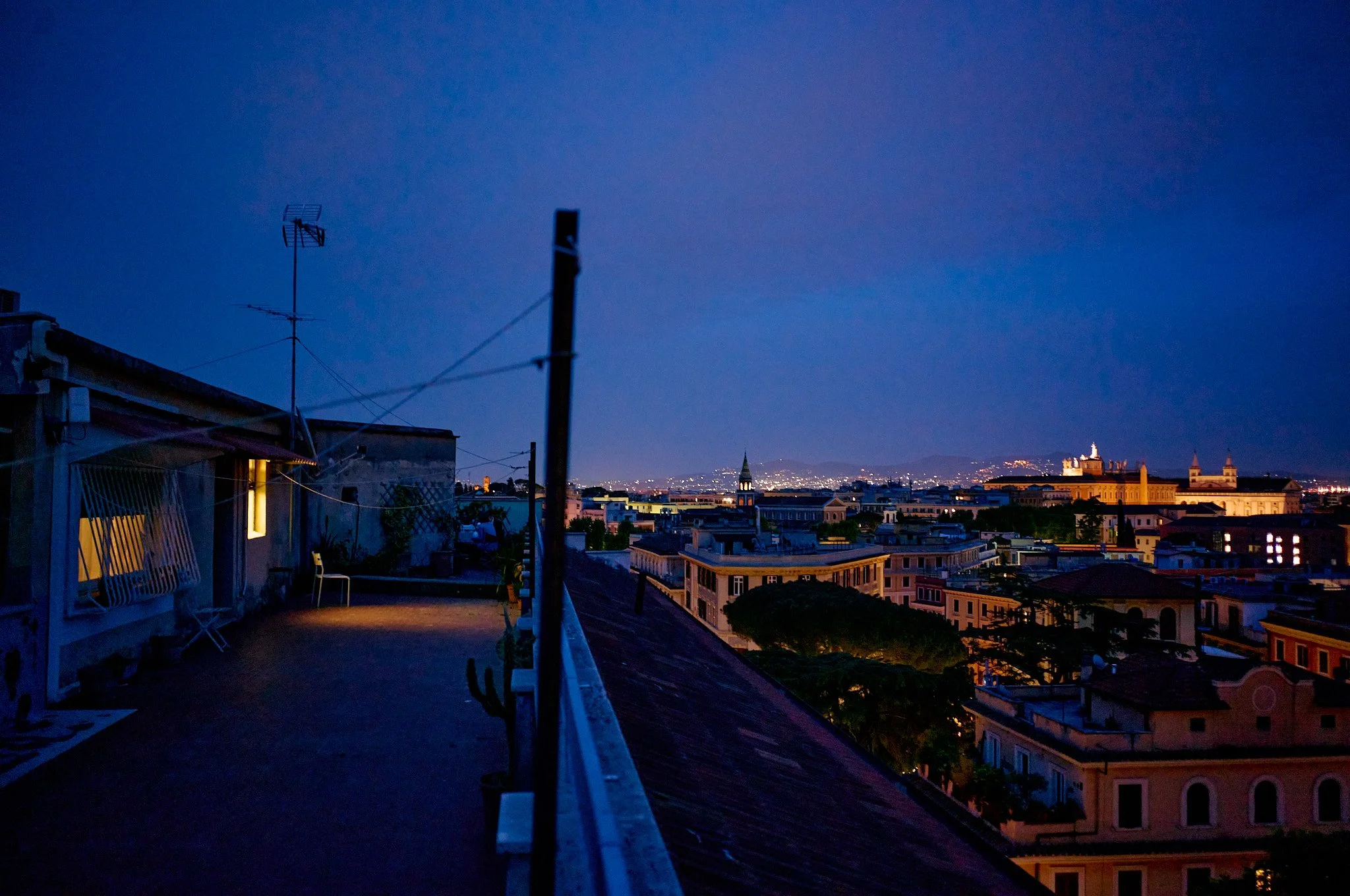 Nighttime cityscape view from a rooftop with outdoor furniture, antennas, and city lights in the distance.