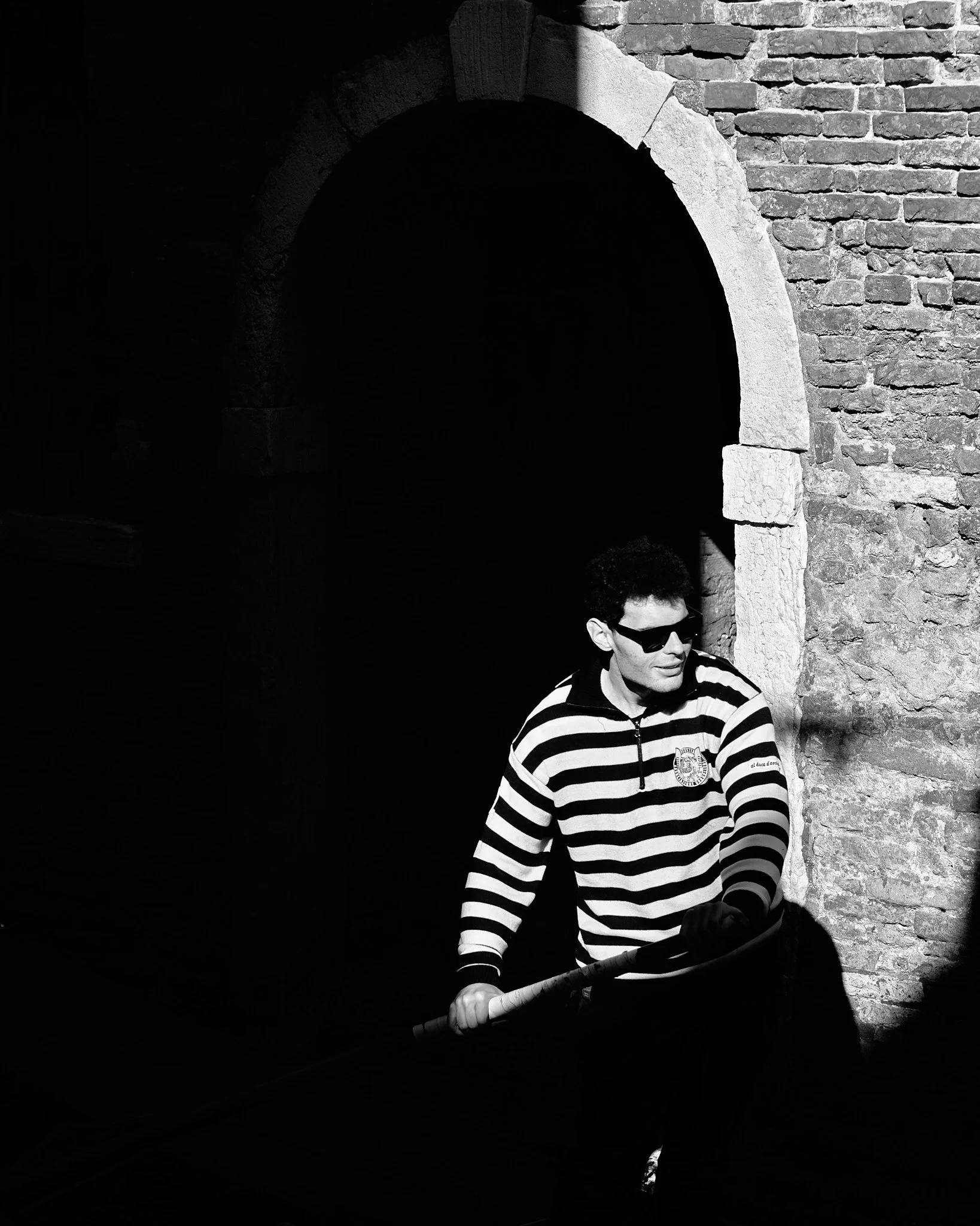 A man wearing sunglasses, a striped shirt, and gloves, holding a stick, standing near a brick wall and archway, with sunlight casting shadows.
