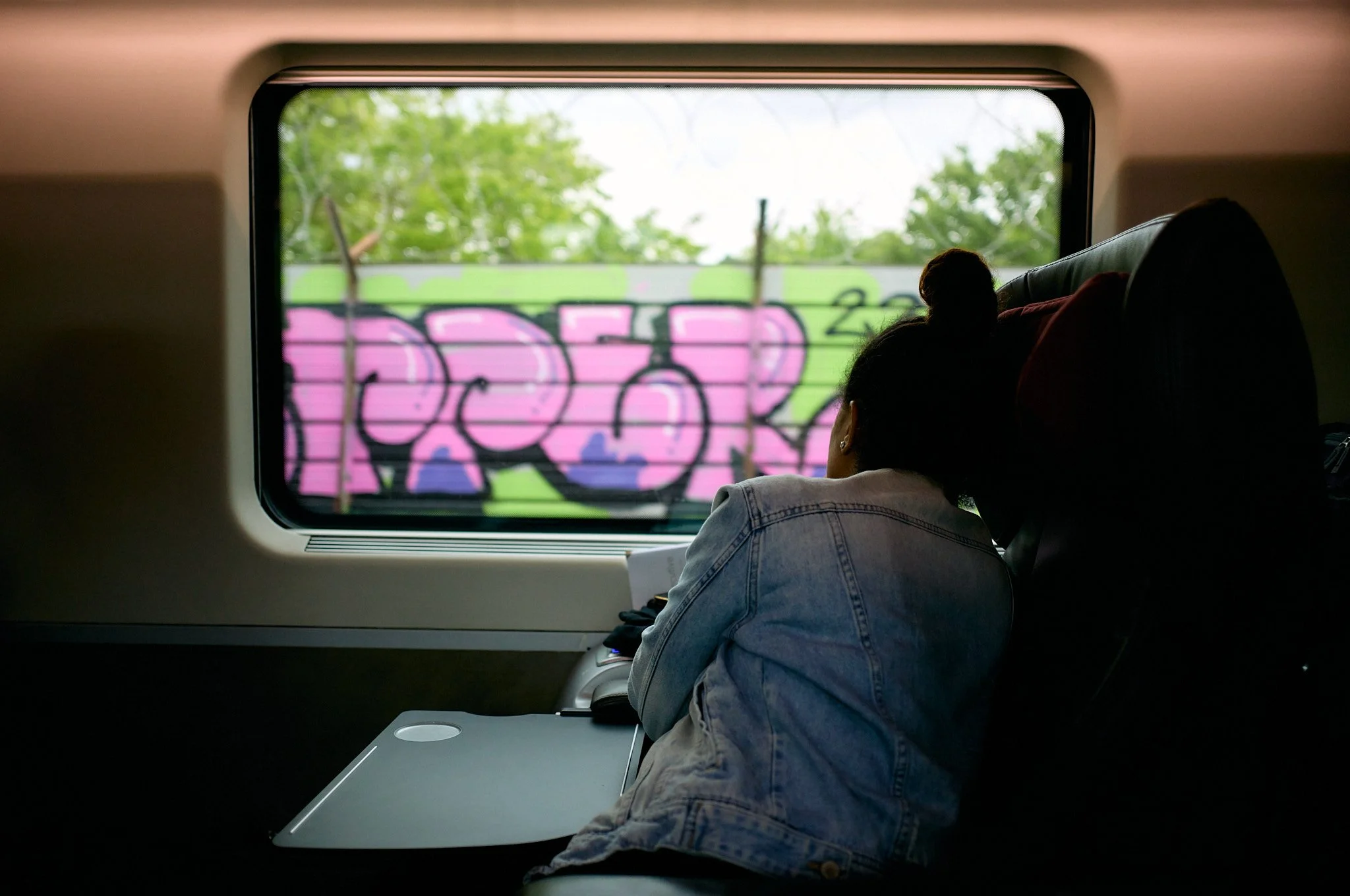 Two women, one with a denim jacket, are sitting on a train looking out the window at pink and purple graffiti on a wall outside.