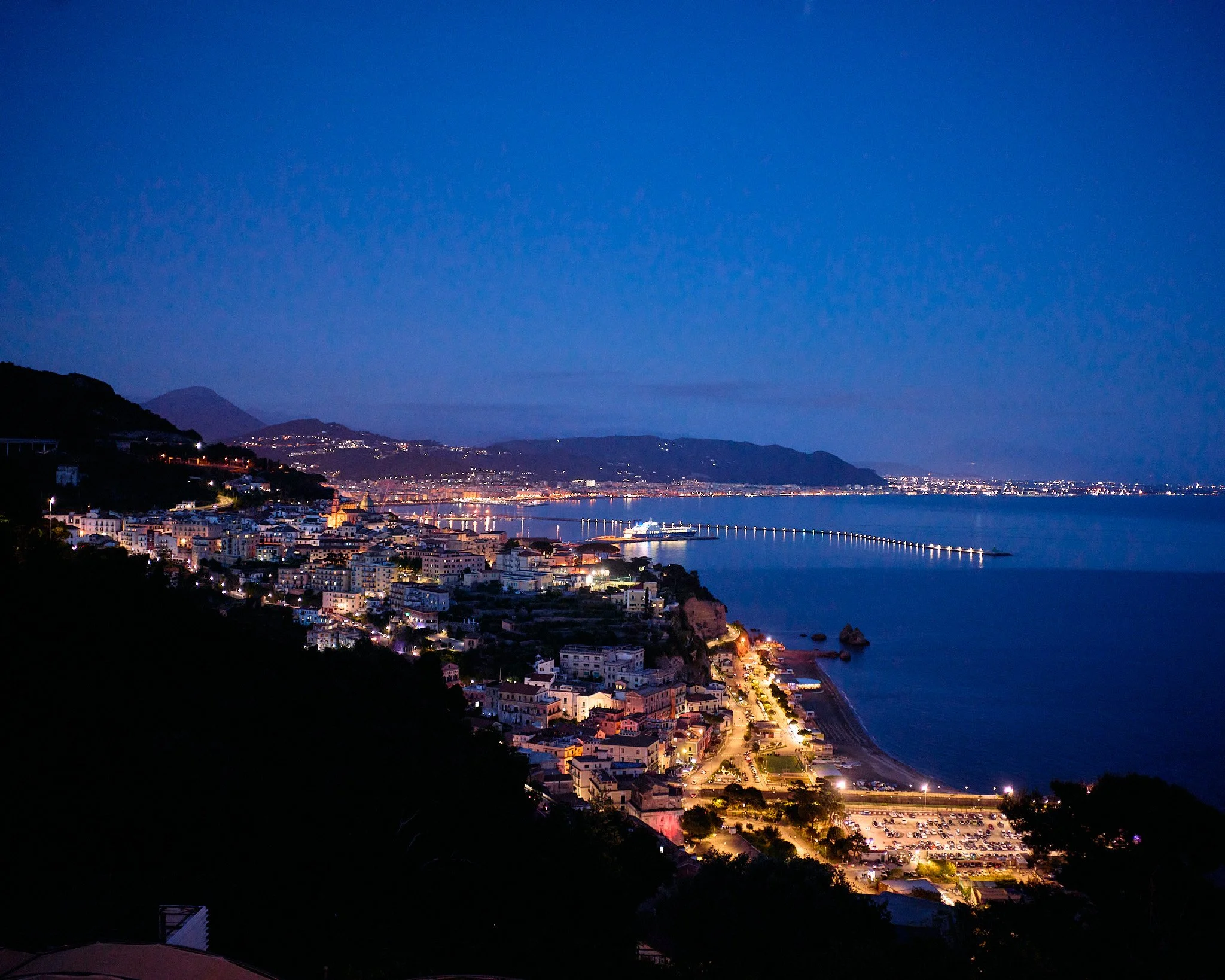 Nighttime view of a coastal city with illuminated streets, buildings, and a pier extending into the ocean, surrounded by hills and mountains.