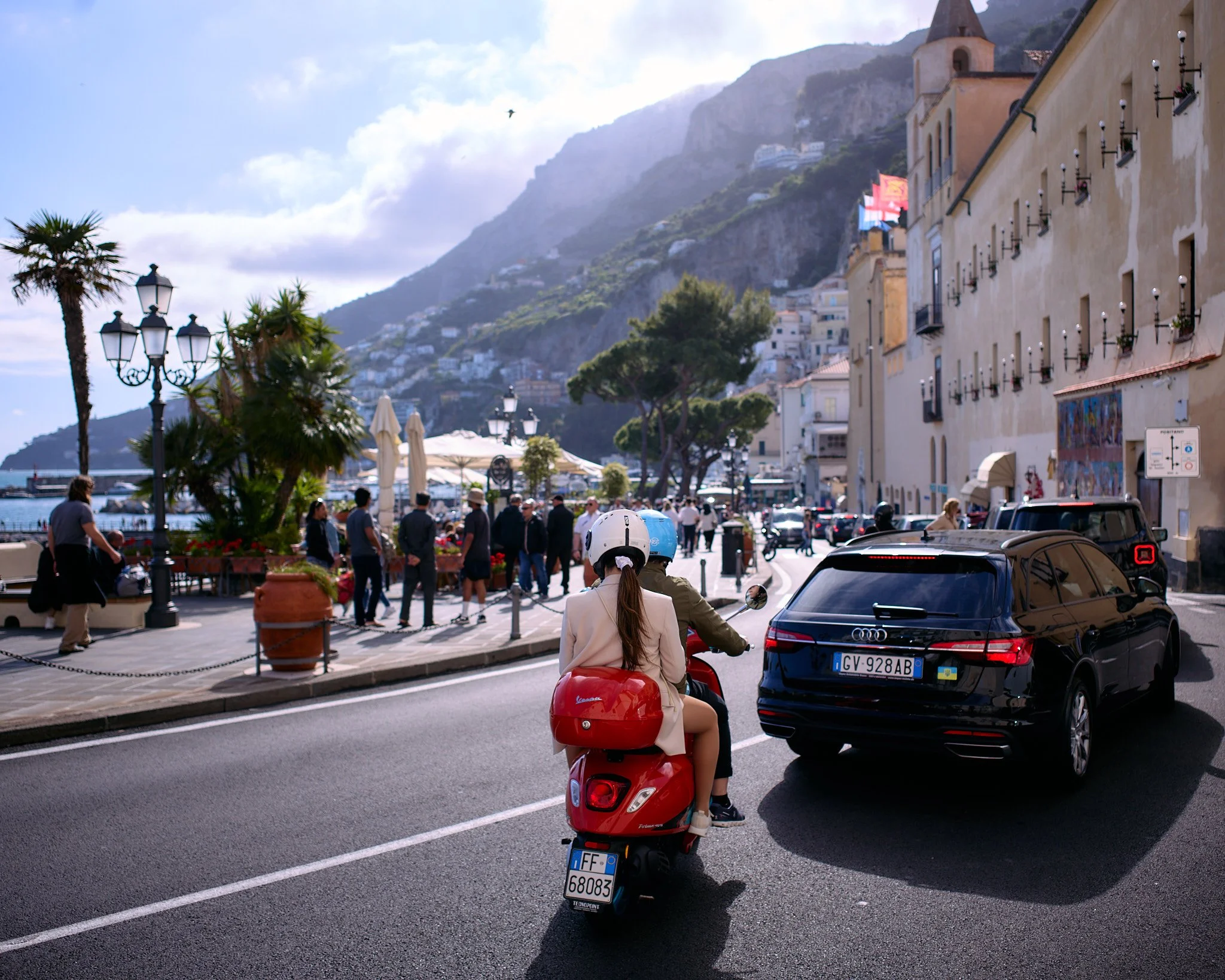 A street scene in a coastal town with people walking and sitting outdoors, a woman and man riding a red scooter, parked cars, a large beige building with windows, and mountains in the background.
